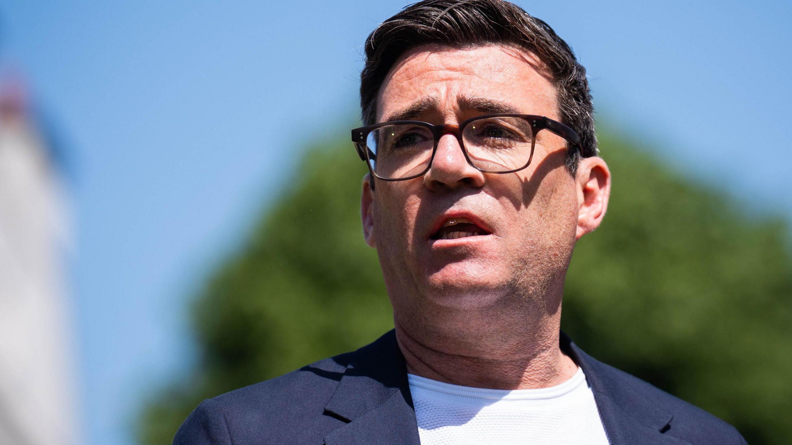 Andy Burnham, with short dark hair and glasses, wears a white T-shirt and blue jacket and stands in front of a blurred image of a green tree