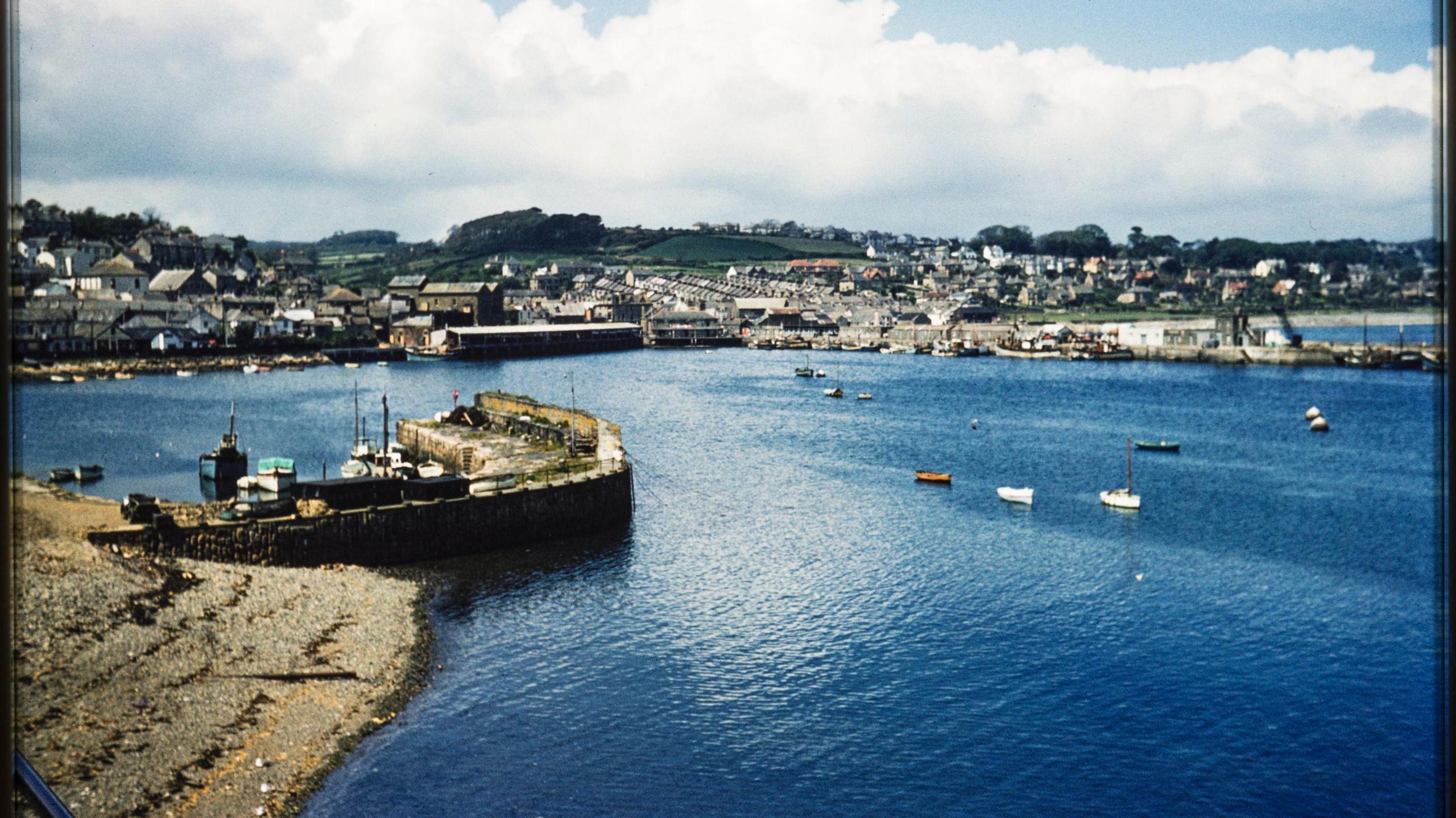 Newlyn's harbour is pictured. It is a small harbour with stone walls. It is surrounded by a large body of blue water.
