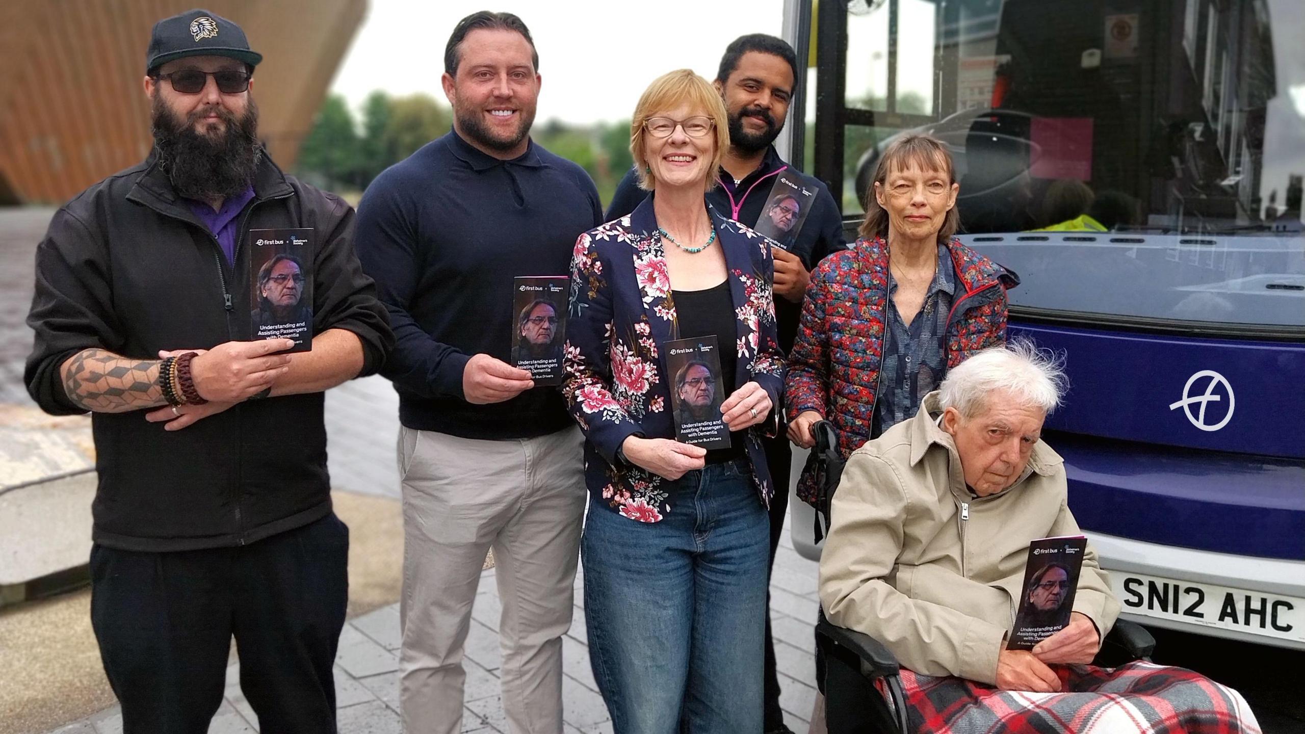 A group of people, including an older man sat in a wheelchair, are stood in front of a bus holding leaflets and smiling.