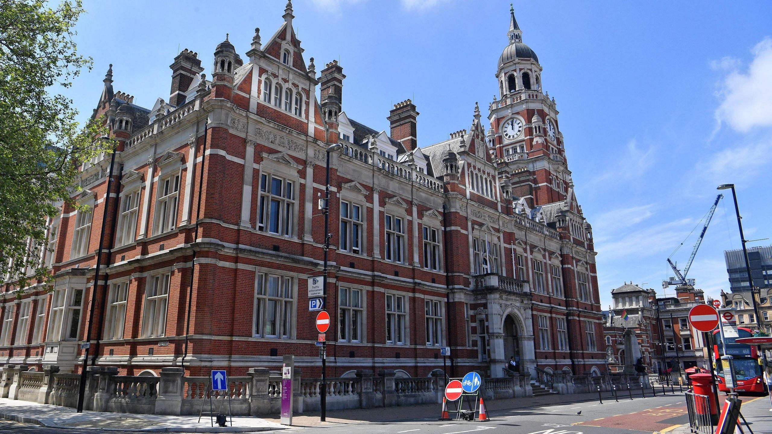 Croydon Town Hall taken from outside, a large red bricked building in the Victorian style with a clock tower and large stone archway entrance.
