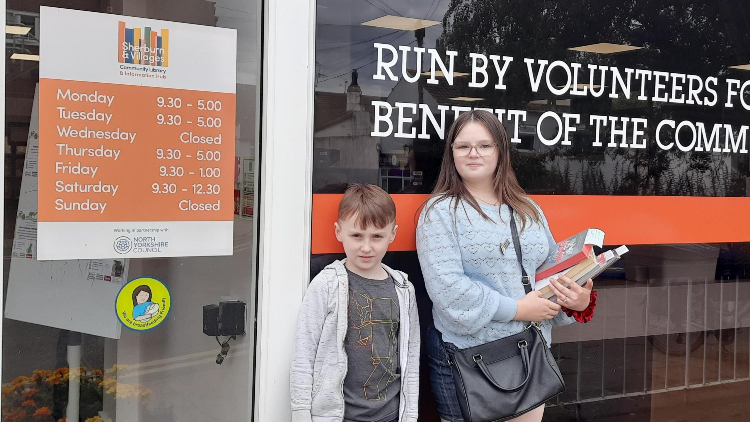 Kaitlin and her brother Toby stand outside Sherburn library. Kaitlin wears a blue jumper and a black handbag and holds several books. Toby wears a grey hoodie.