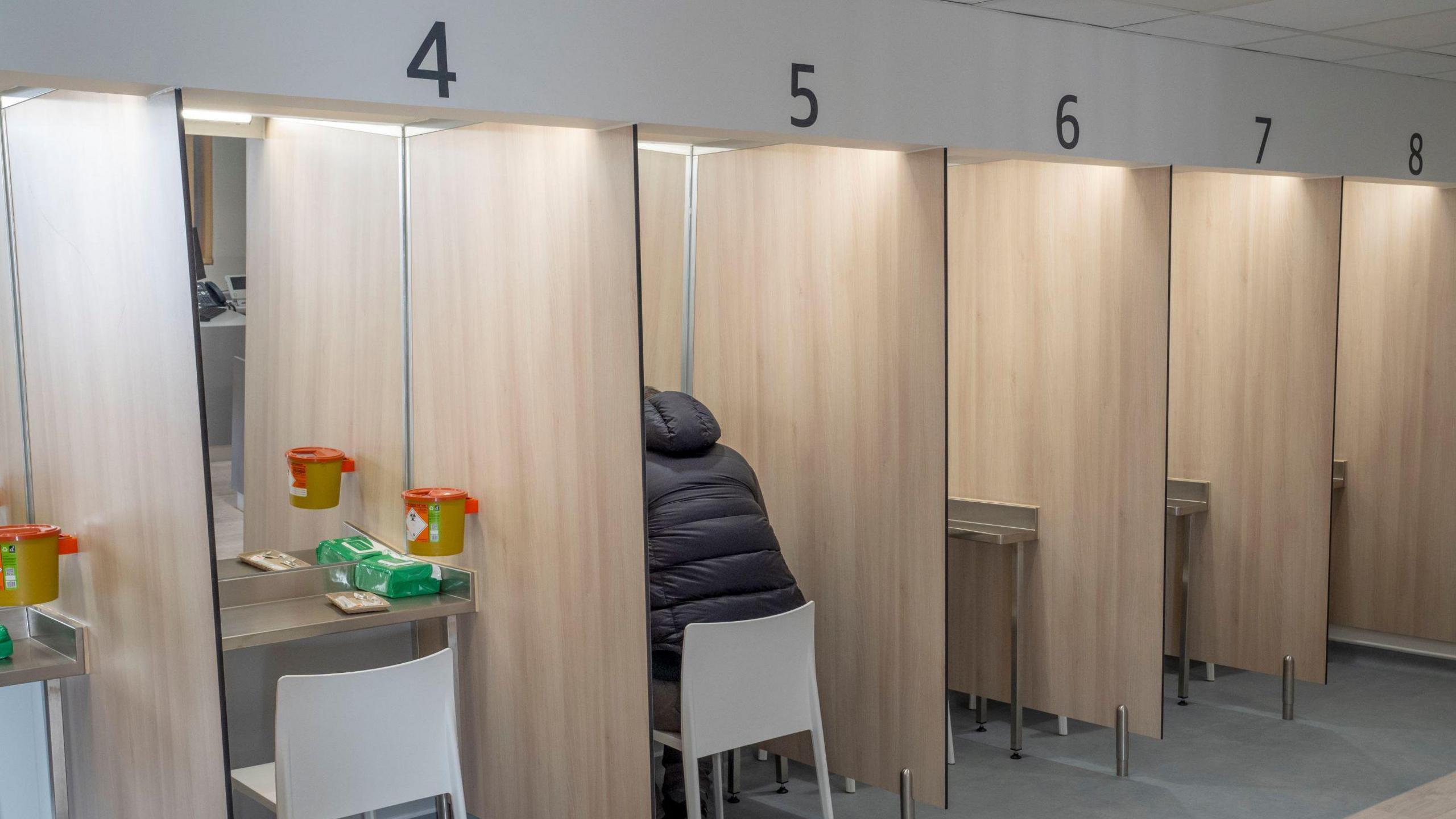 Several cubicles, numbered from 4 to 8, lined up - there are mirrors at each one, with a chair and other items on each table. A man wearing a dark jacket, with his head not visible is sitting at No 5.