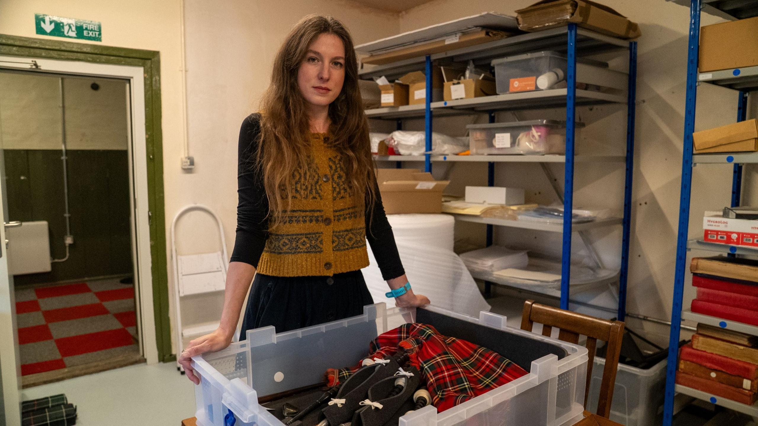 Susan Higgins, curator at the King's Own Scottish Borderers museum in Berwick-upon-Tweed, a long-haired young woman in a knitted waistcoat, photographed with WW1 piper Daniel Laidlaw's bagpipes - they are in what looks like a red Stewart tartan and are kept in a plastic archive box.