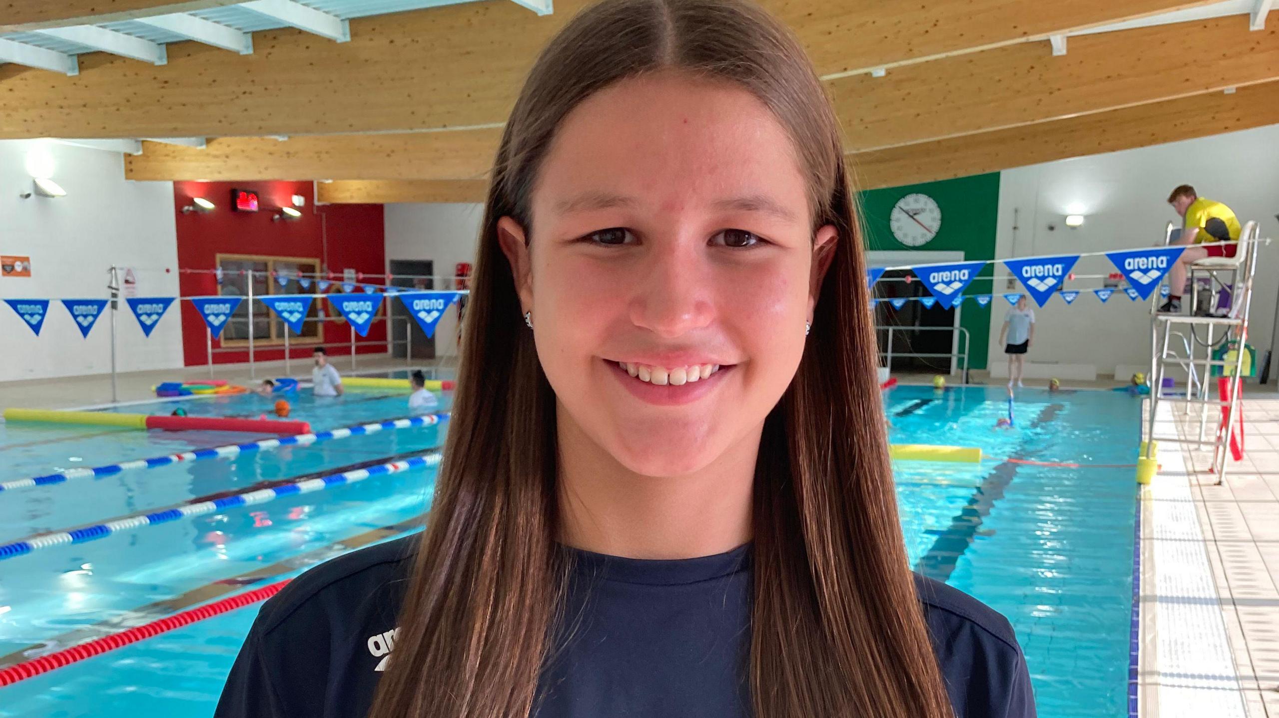 A girl with long light brown hair over her shoulders, wearing a blue T-shirt and standing in front of a bright blue swimming pool, with a life guard on a tall chair to her right and some swimmers to her left