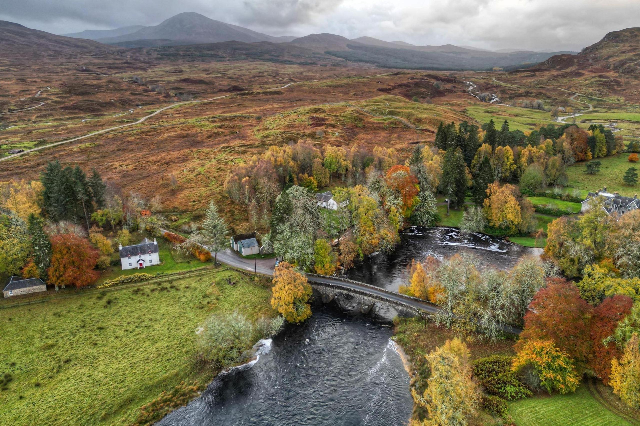 An aerial view of Bridge of Gaur showing an upland landscape of hills and, in the distance, mountains. A stone bridge spans a river and there is a two-storey white walled house among tree on the riverside.