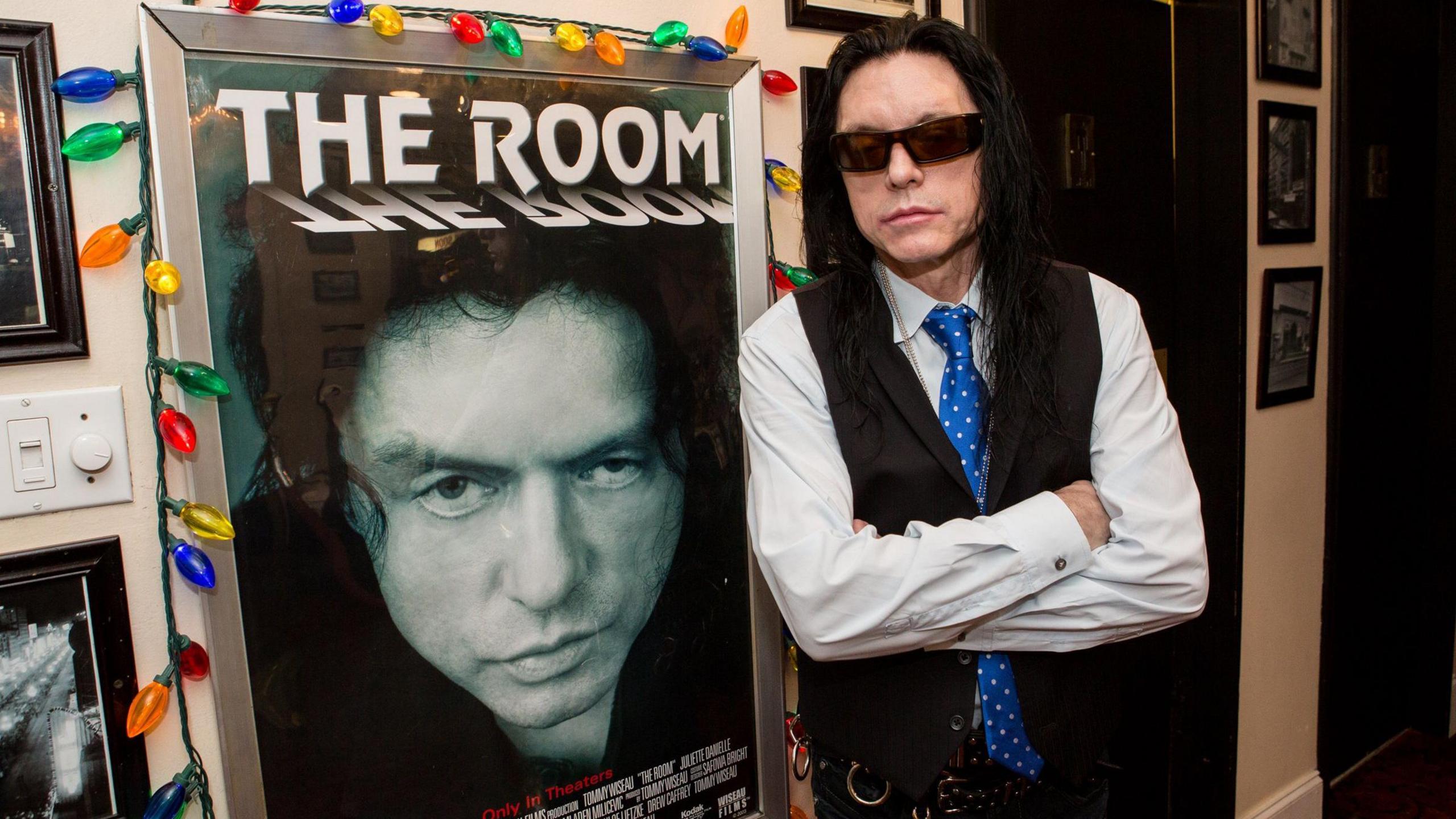 Tommy Wiseau with long black hair, black glasses and a black waistcoat leans against a poster for The Room, framed by coloured fairy lights, at a screening in New Orleans in 2014