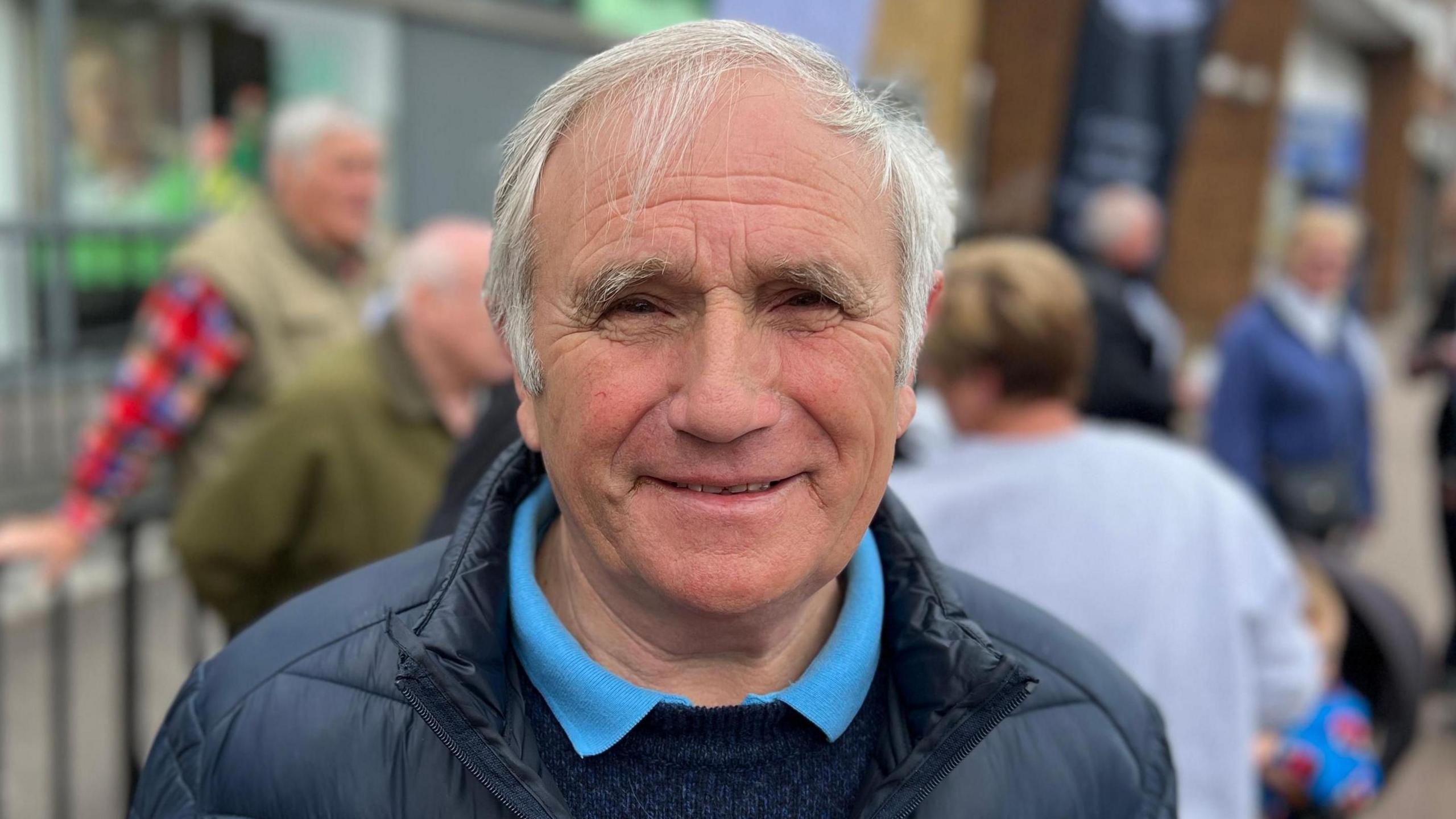 Dave Blackwell, who has grey hair and is wearing a navy coat, with a navy jumper and blue polo top underneath. He is smiling at the camera while standing in front of other people, who have been blurred out.