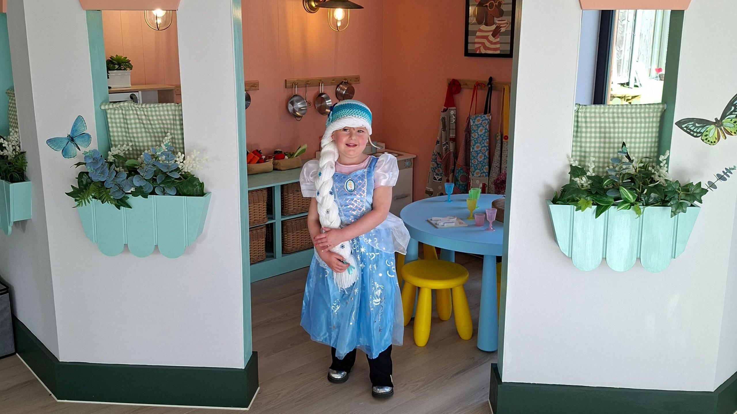 A five-year-old girl is wearing a blue dress and cap with a white plaited hair extension stetching to her waist. She is standing in a child's house with a table and chair and kitchen items in it. It has faux flower troughs underneath faux windows.