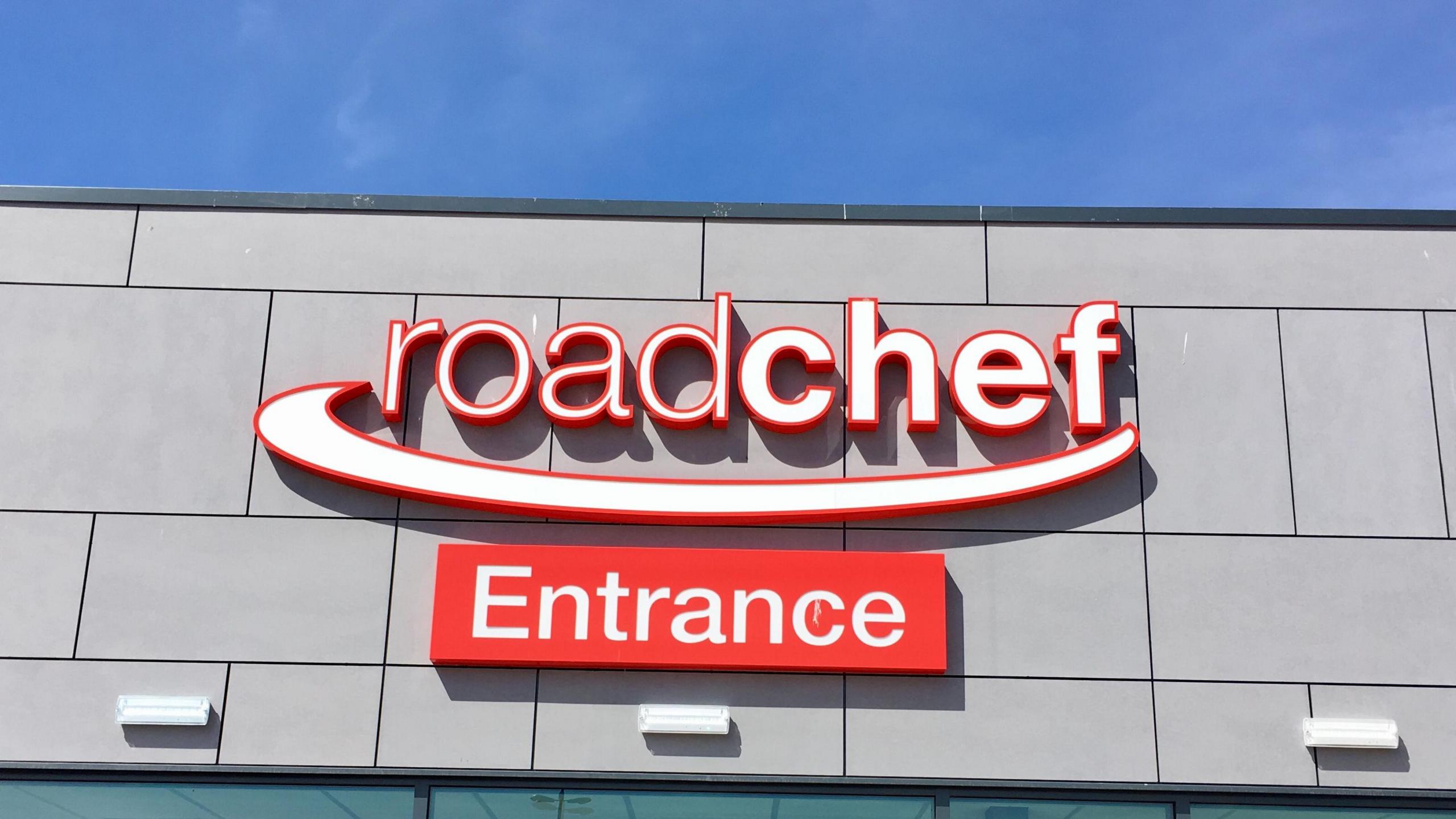 Front view of a red and white Roadchef sign. An additional sign that reads "Entrance" is mounted below.