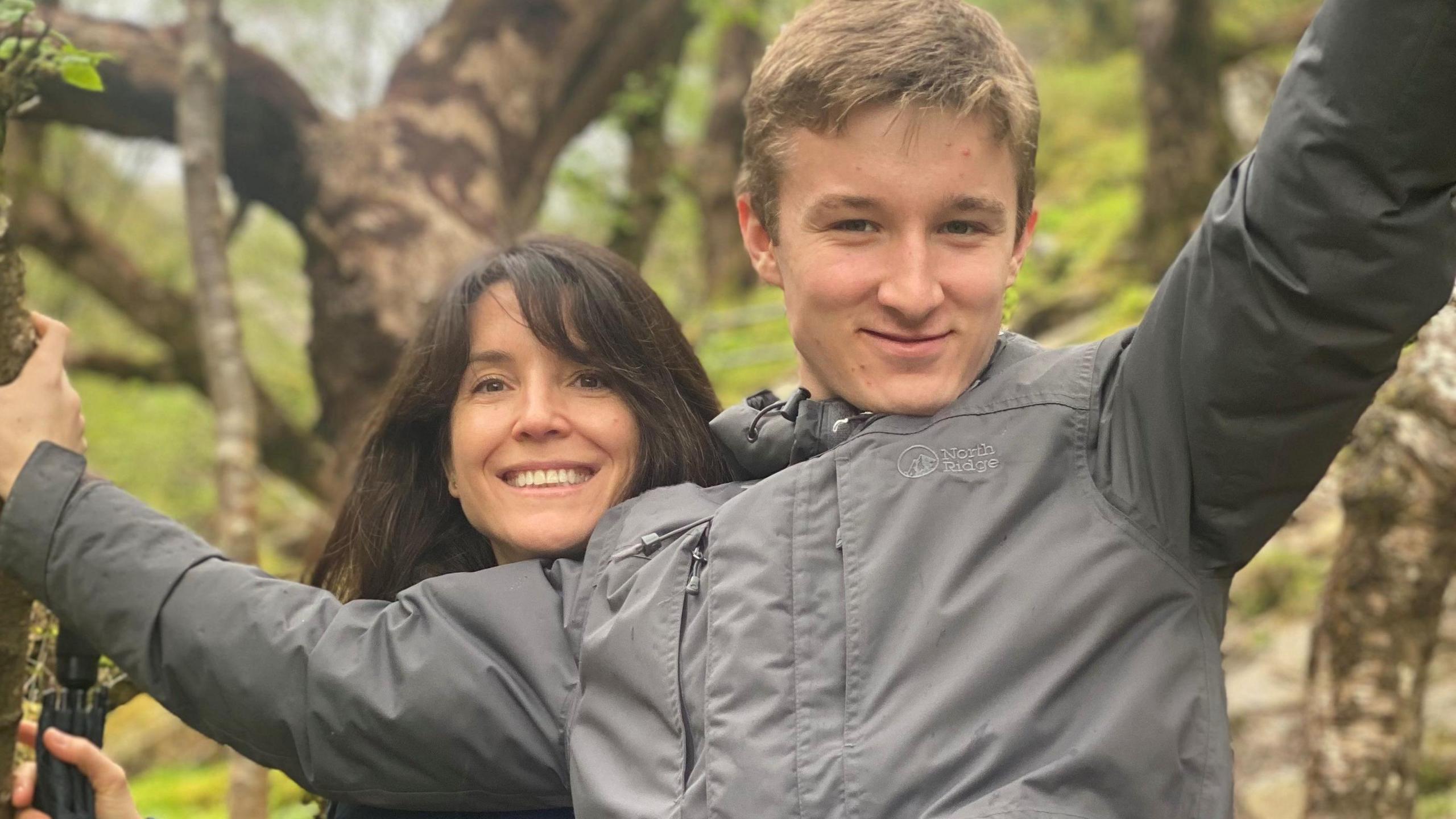 James, smiling wearing a grey coat with his arms holding on to the branches of a tree. His mother, with long brown hair, is standing behind him with her head resting on his shoulder.