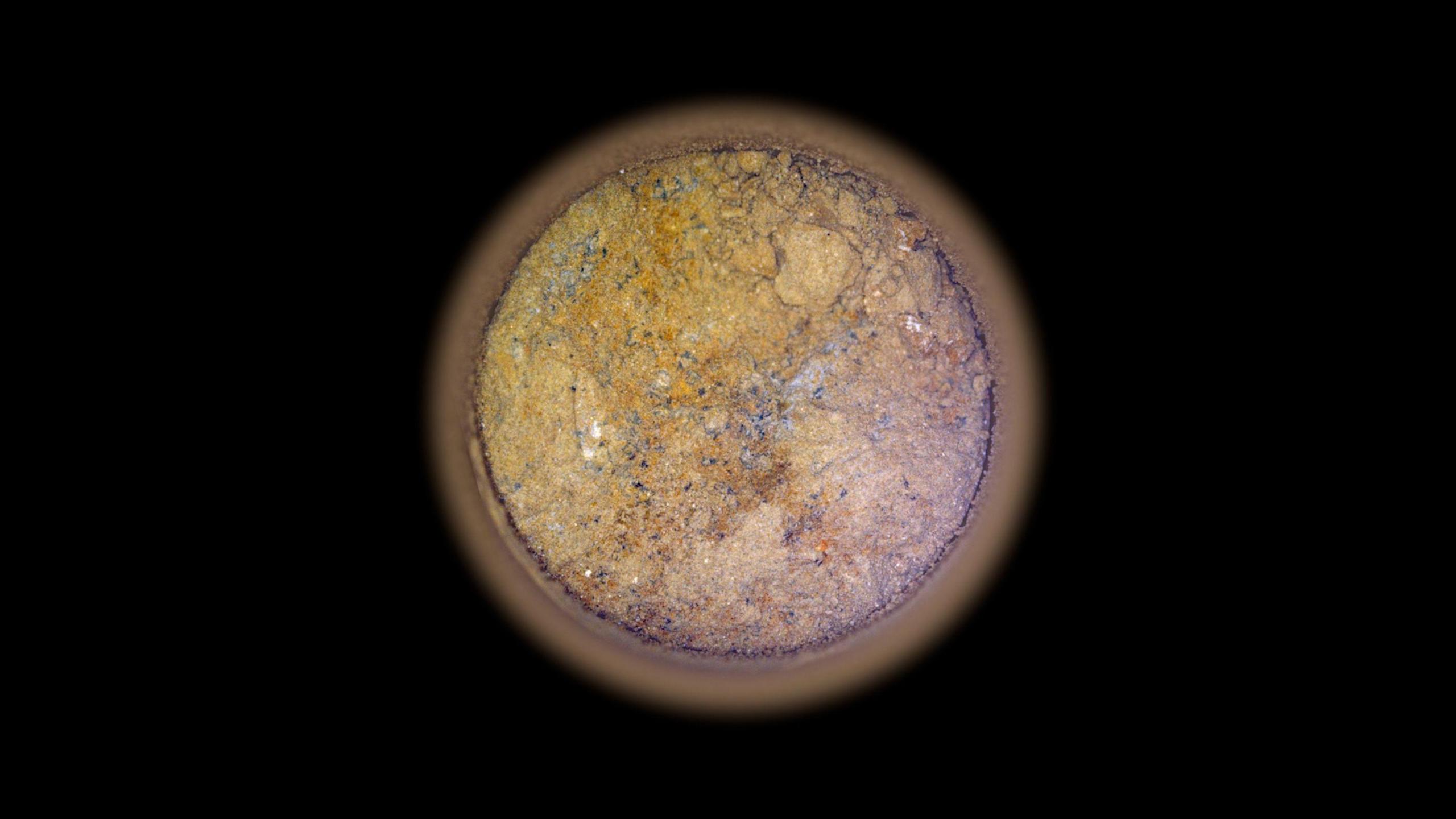 Image of a core of rock taken from Mars. The image shows a circular slice through of the rock sample against a black background. The sample is orange and dotted with white, grey and black marks.