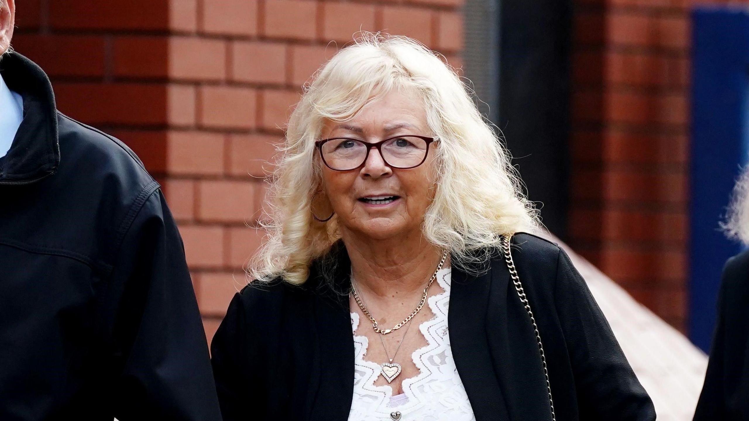 Karen Spragg with light, shoulder-length hair and black-rimmed glasses, in front of a red-brick building.