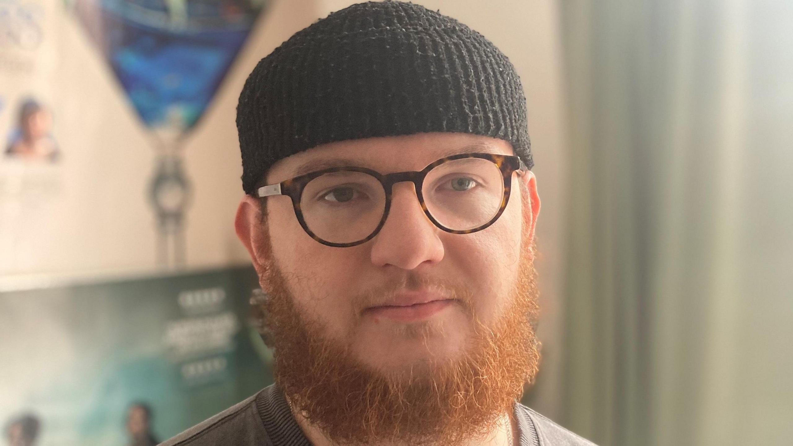 A man wearing a black hat faces the camera. He has a beard and is wearing glasses. 