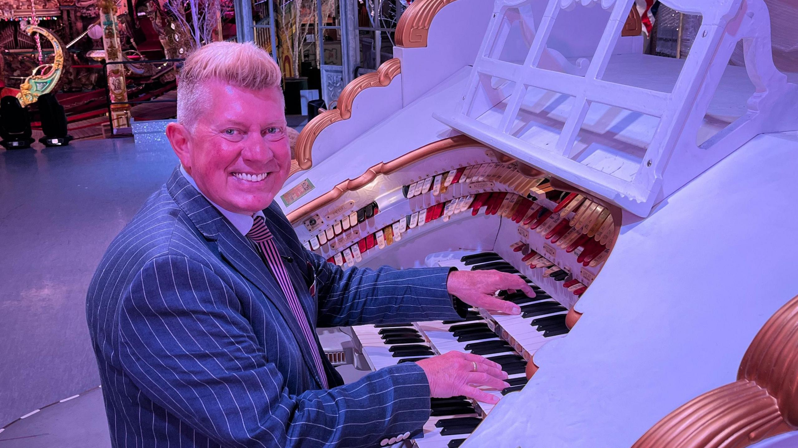 Robert Wolfe is sat on a stool on the left playing the Wurlitzer which is white with gold accents. He is looking towards the camera smiling, and is wearing a navy pinstripe suit jacket. 