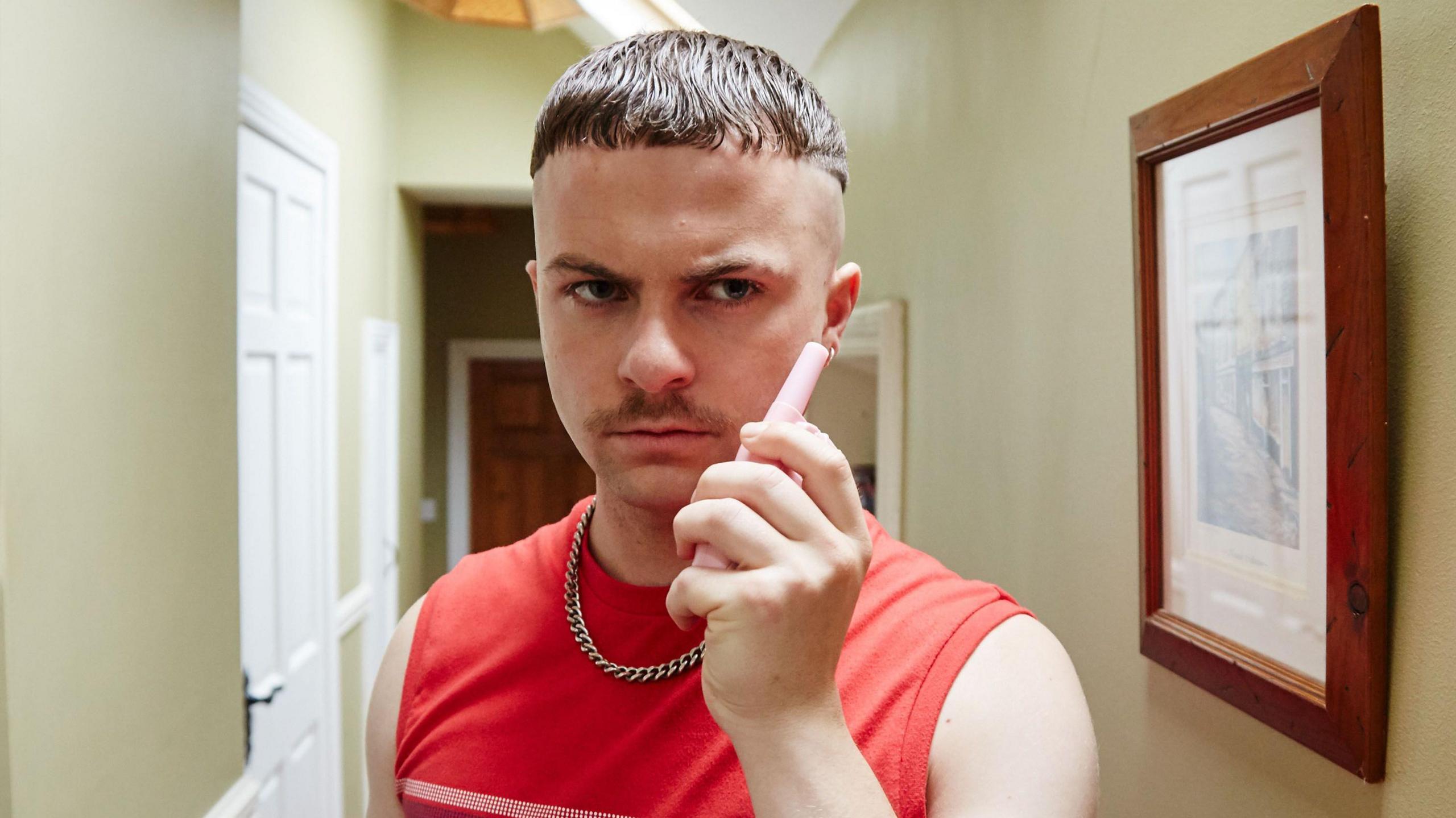 Alex Murphy as Conor MacSweeney in The Young Offenders. He is standing in the middle of a beige hallway wearing a red vest top and a silver chain necklace. He is holding a pink walkie talkie device in his hand.