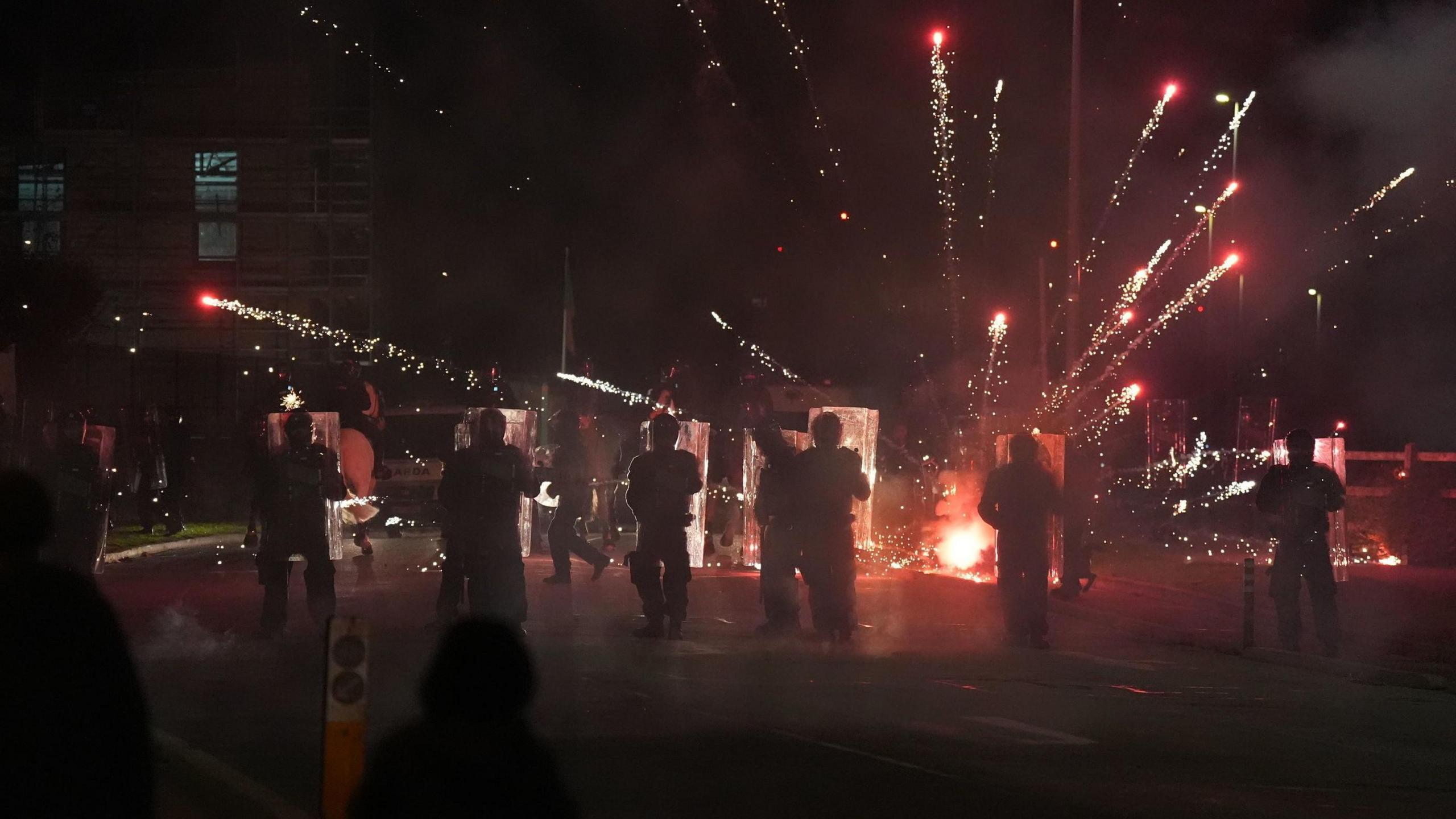 A street at night-time. The backs of silhouetted riot police, holding transparent riot shields, can be seen from behind. In front of them, a firework has just exploded, sending a number of red sparks shooting in all directions. In the foreground the silhouettes of a few onlookers can be seen.