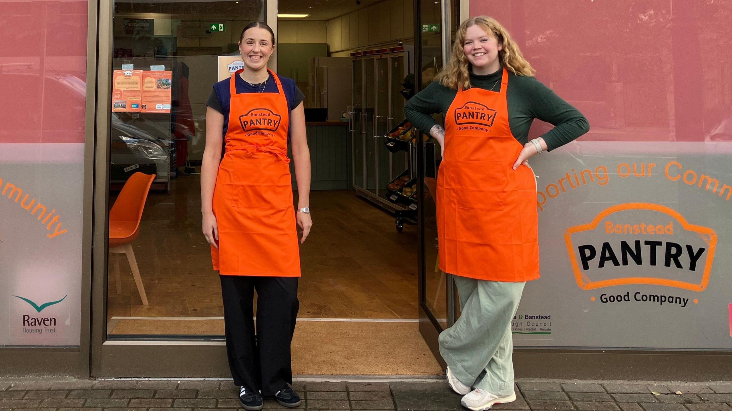 Two women in orange aprons pose outside a shop