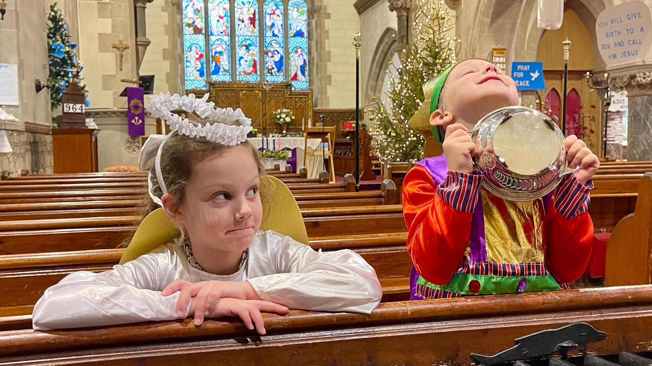 Two children sit on a pew in a church. The girl on the left is wearing an angel outfit complete with a halo headband. She is staring at the boy on the right dressed as he king. He is wearing a crown and a gown of green, gold, red and purple. He is holding a metallic dish to his chest and looking at the ceiling. Christmas trees and flowers can be seen at the alter.