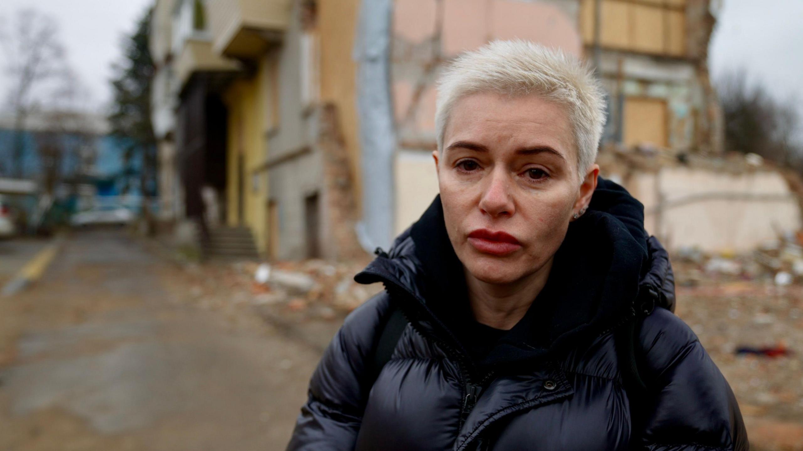 A lady with short blonde hair looks into the camera. Behind her is her apartment which has been totally demolished by a bomb.