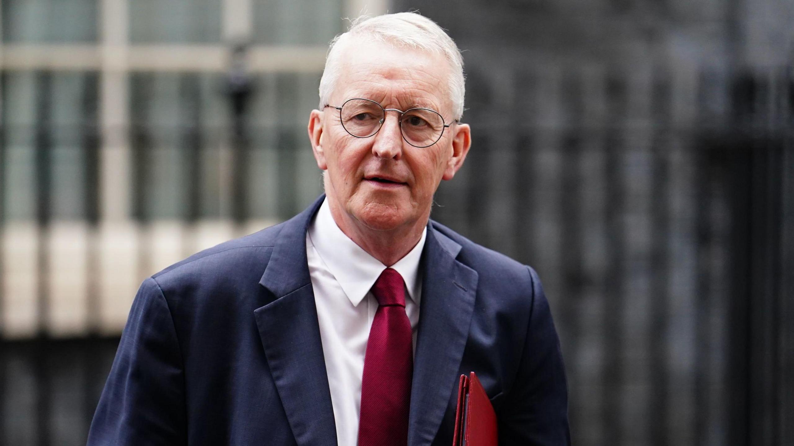 Hilary Benn is an older man with short white hair. He has a pair of thin, circular glasses and is walking outside a grey brick building with a black iron gate in front. He is wearing a navy suit jacket, a white collared shirt and a red tie. He is also carrying a red leather folder.