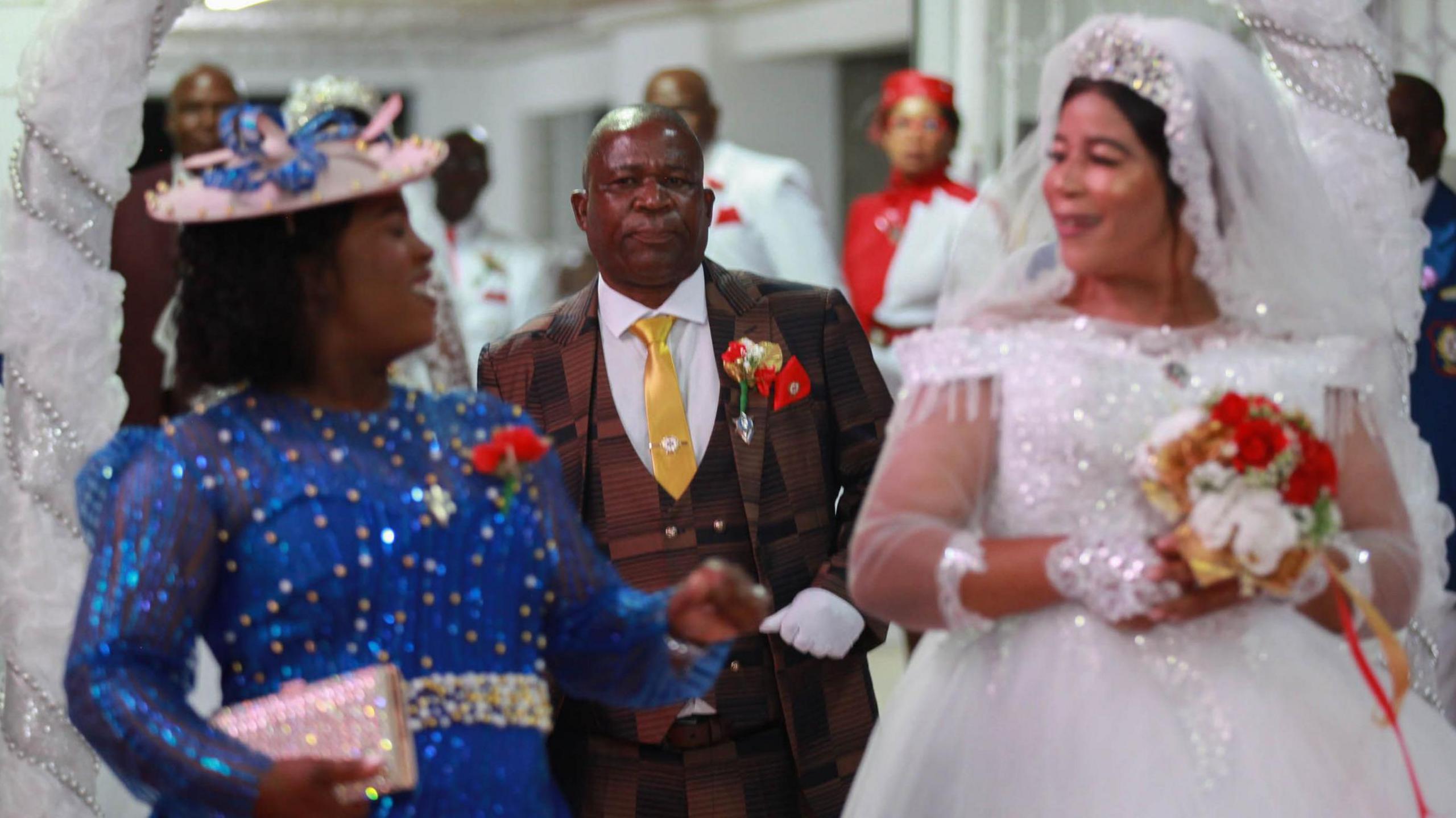 Shirley Molala (L) in a sequin blue dress and pink hat laughs as she looks at Evelyn Sekgalakane, who smiles. She is wearing a white bridal gown and veil and holding a bouquet of red and white flowers. Behind them is Lesiba Molala in a brown patterned suit, yellow tie and white shirt.