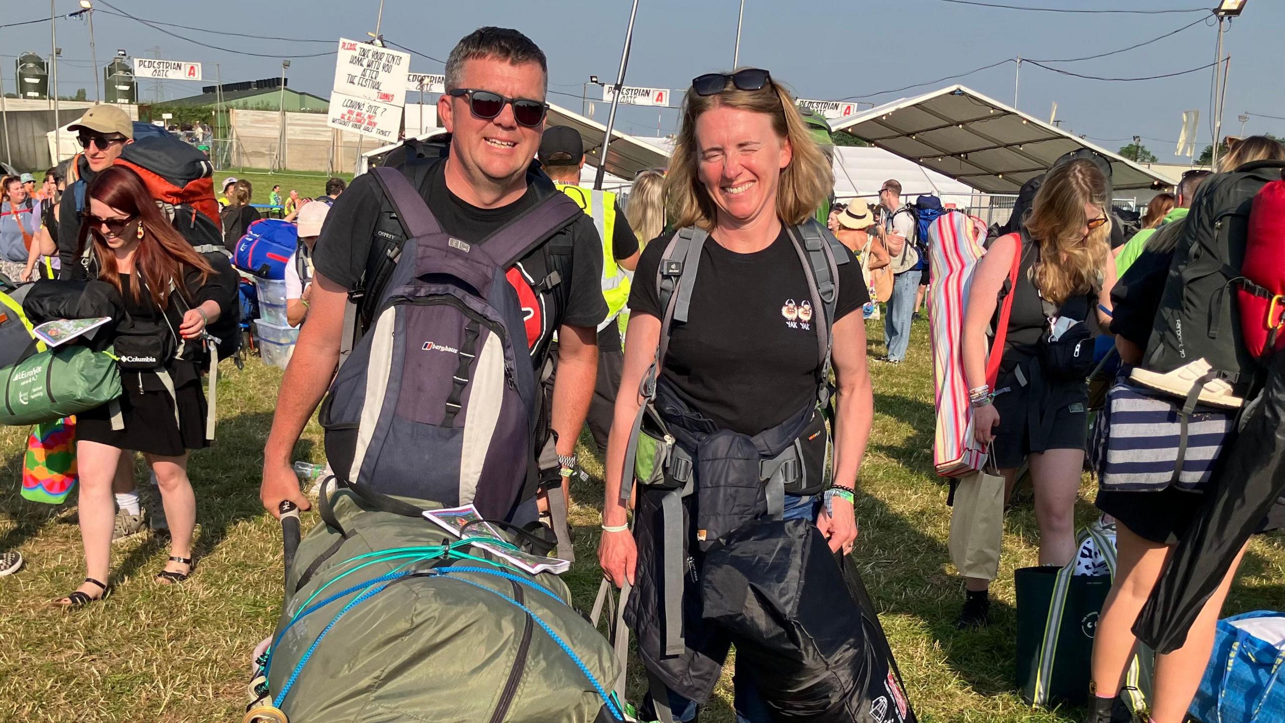 Tom and Emma at the entrance to the site holding backpacks and a large wheelbarrow. 