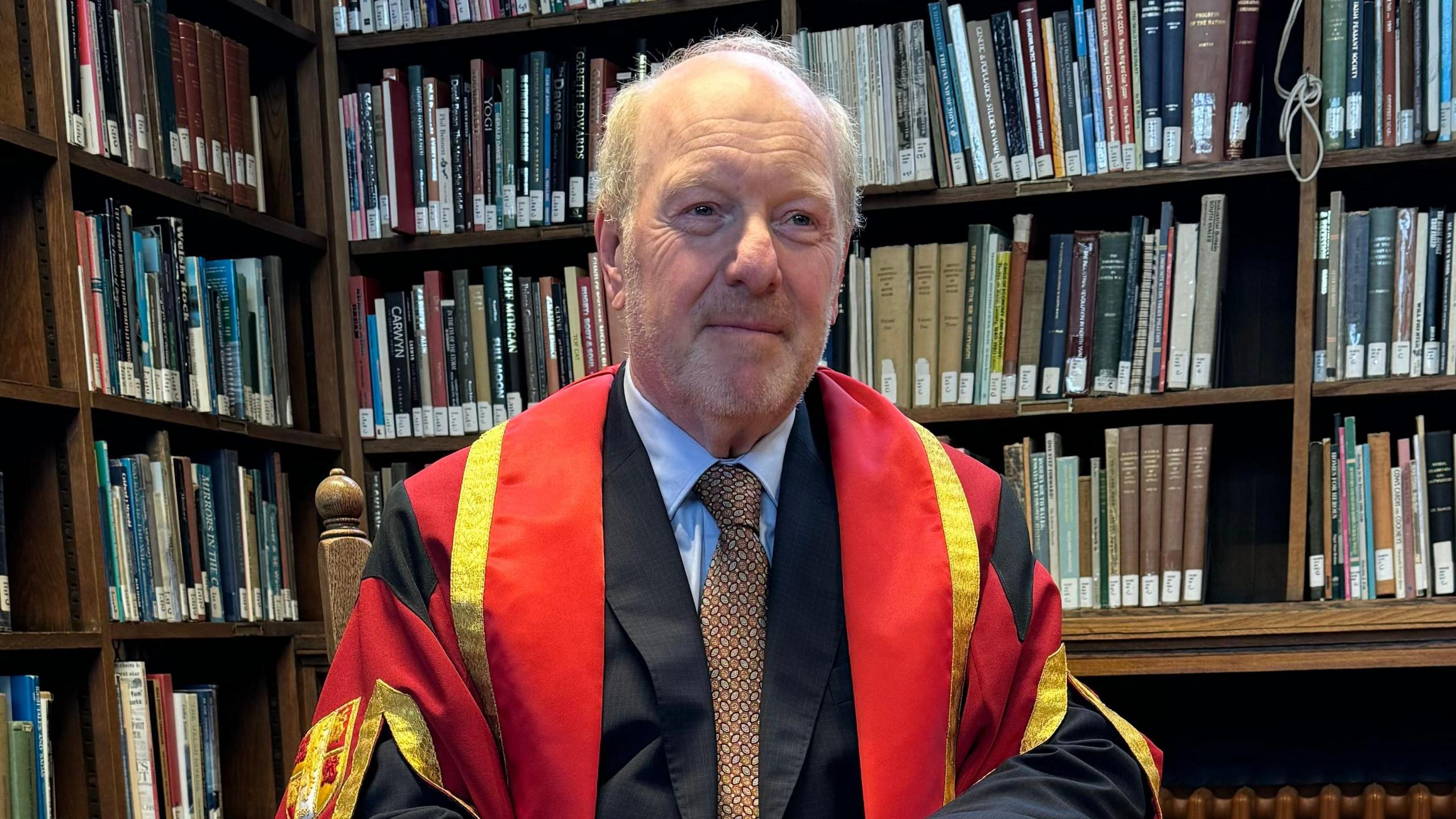 Sir Alan Bates sits in Bangor University library, wearing red, gold and black doctorate gown and a black suit, with a blue shirt and brown tie. Behind him are shelves of books.