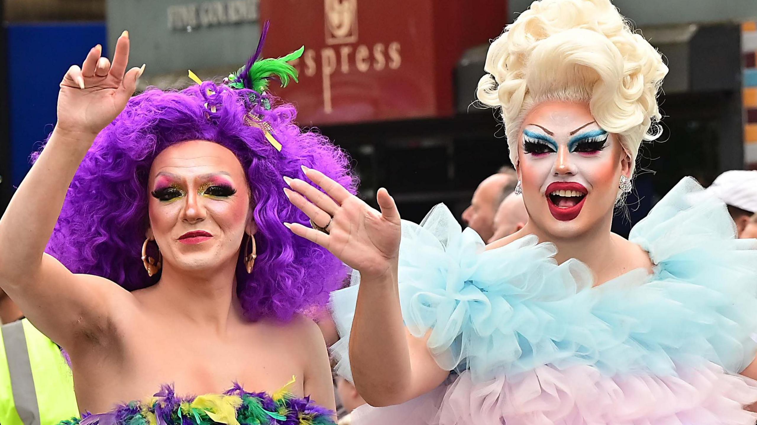 Two people dressed up. One is wearing a purple wig with flowers and the other is wearing a blond wig. They're both wearing colourful make-up.