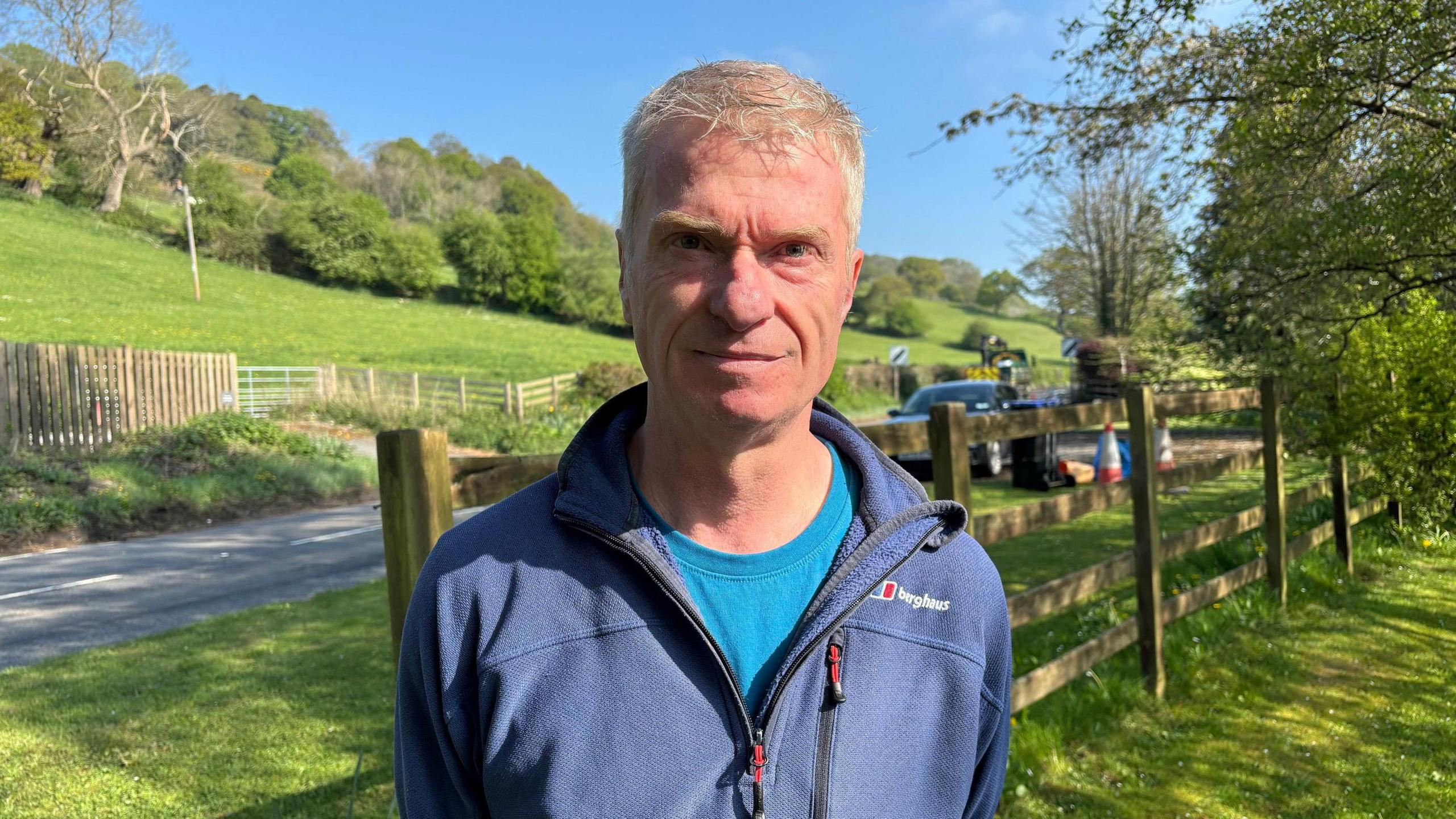 Tony Johnson at the scene behind damaged fence on the grass next to the road. He is wearing a light blue t-shirt under a blue zipped jumper and has short blonde hair