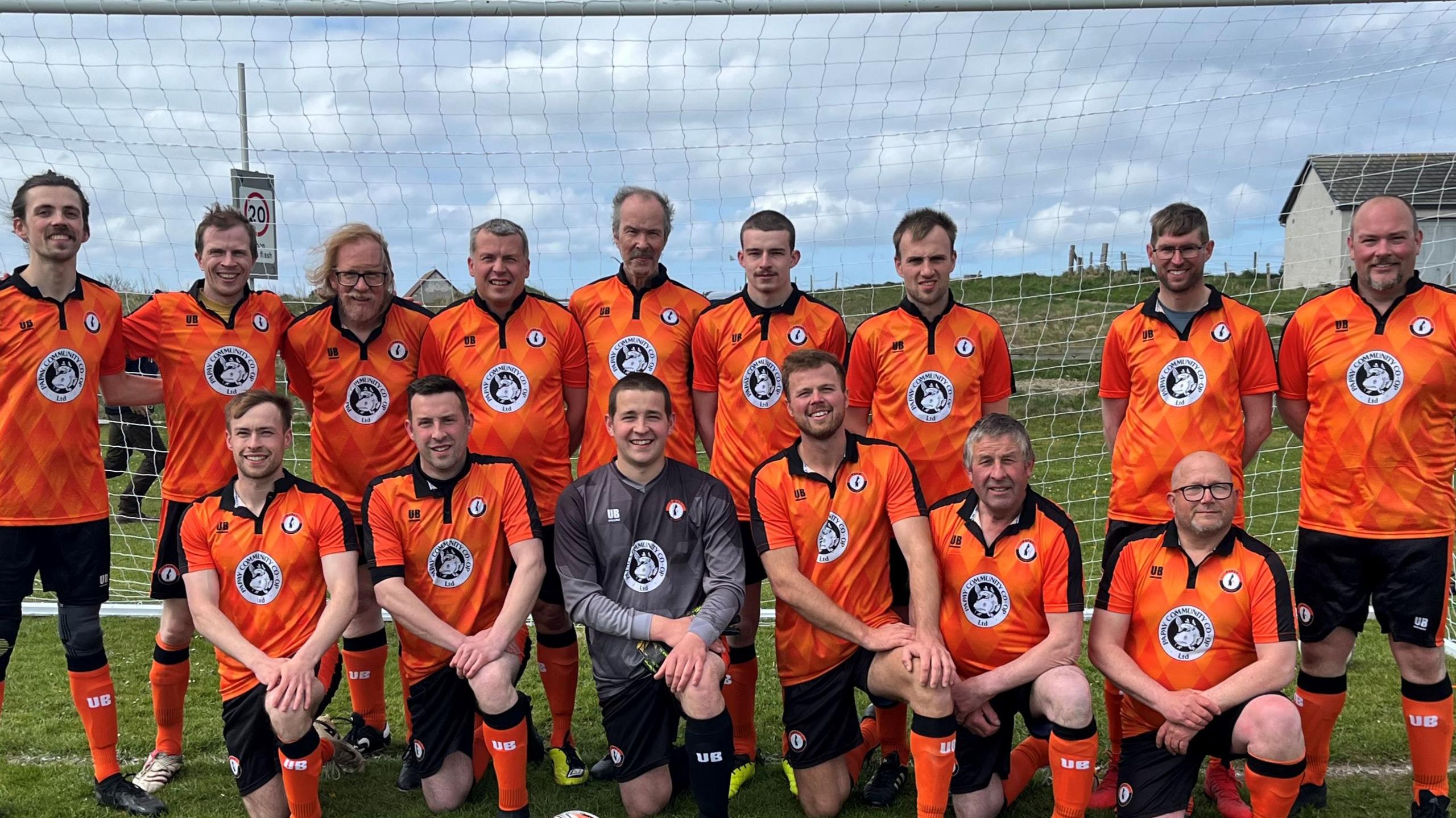 Two rows of football players in orange tops standing in front of goalposts.