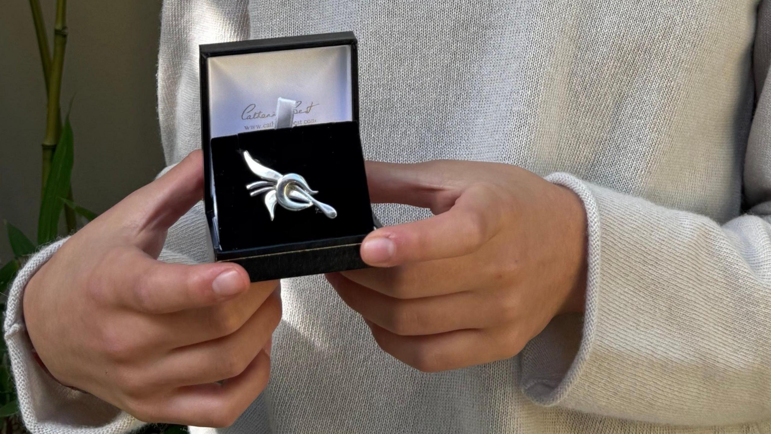 Juliette's hands holding a black jewellery-type case which is open, showing an intricate silver brooch lying on a black velvet cushion.