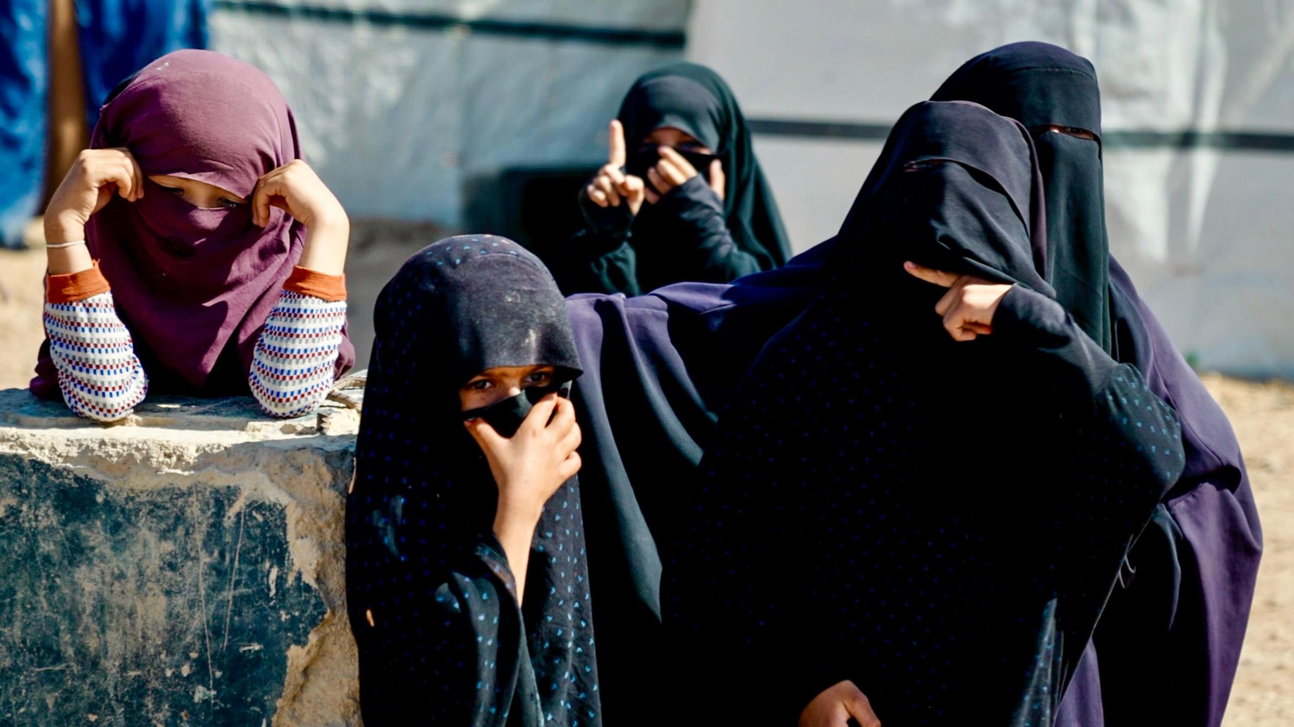 Veiled women, some are clad head to toe in black. Thee are also a few children. One is raising her index finger. A woman is making a cutting gesture.
