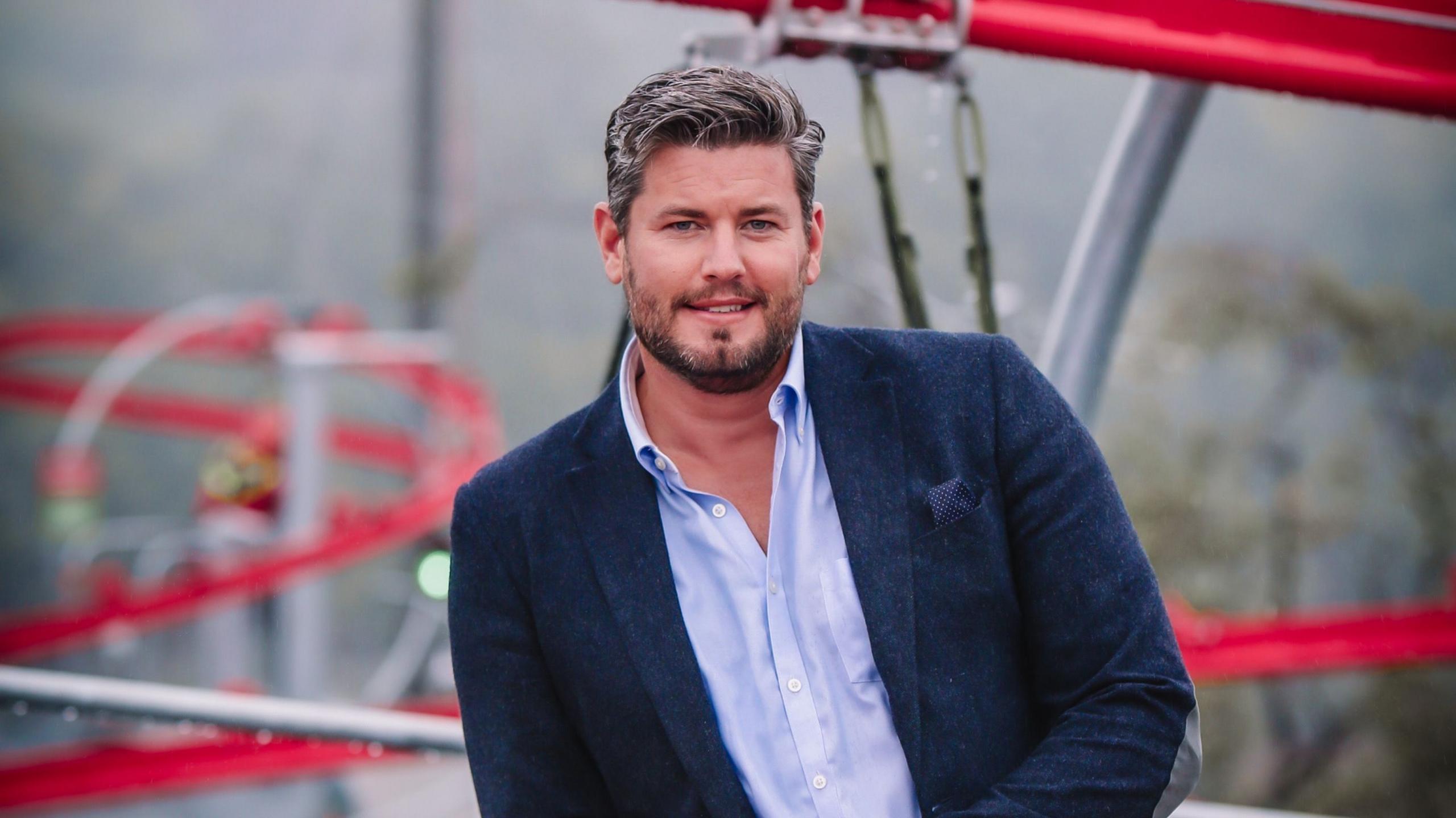 Andrew Hudson, a bearded man in open-neck shirt and blue blazer. In the background, out of focus, are the red tracks for a roller-coaster ride.