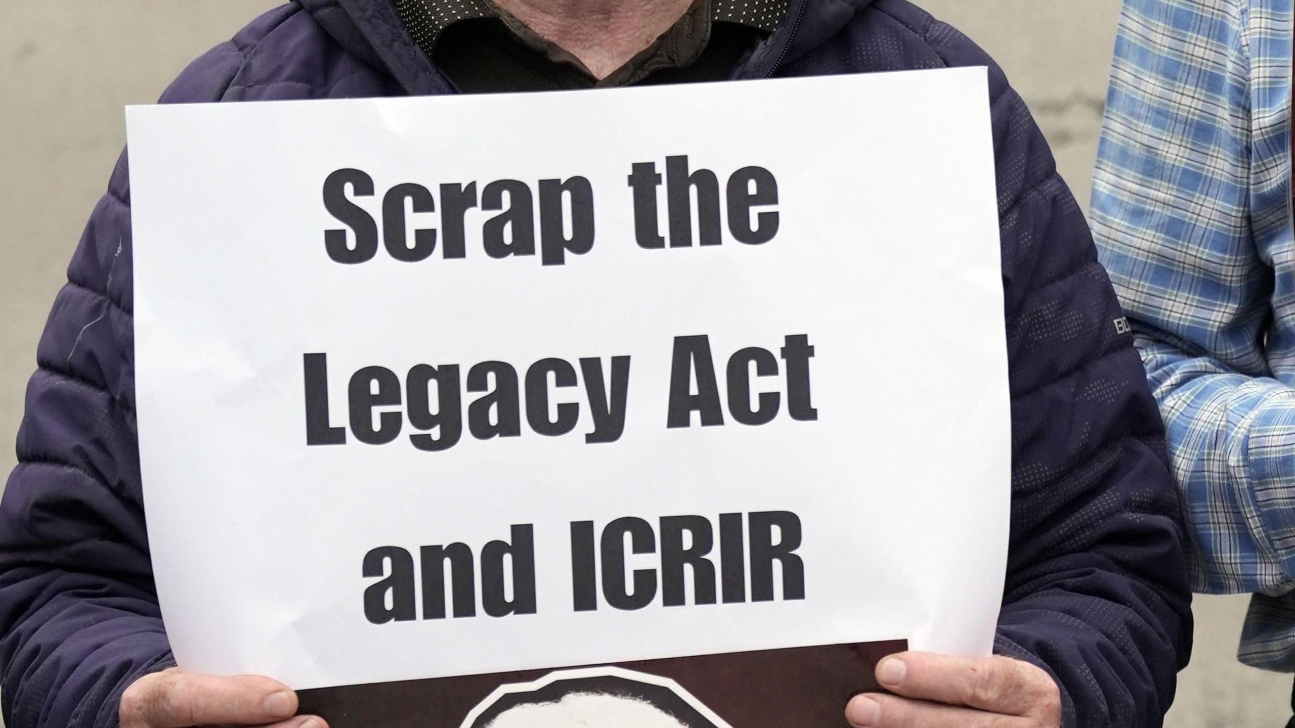 A large sign is printed on white paper reading 'scrap the legacy act and I.C.R.I.R' in black text. It is being held by someone in a navy blue coat. Only their fingers are visible on the bottom of the sign, and part of a blue checked shirt worn by someone standing beside them. 