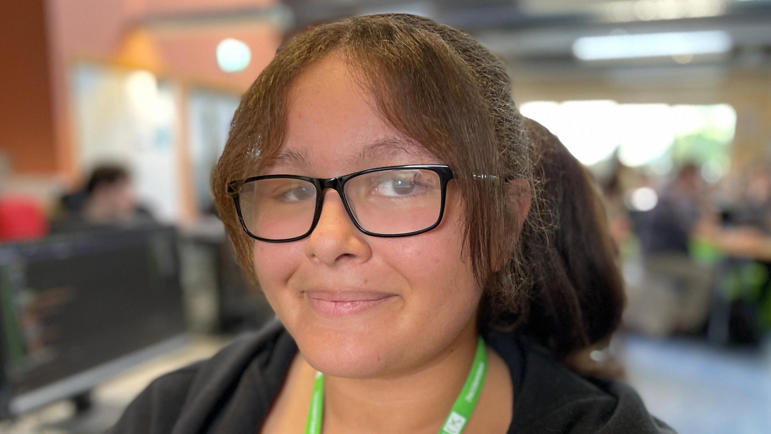 Madi is 17 years old and wears glasses and a green college lanyard. She is wearing a black hoodie and sat in front of a computer screen in a classroom. She is looking directly at the camera and smiling.