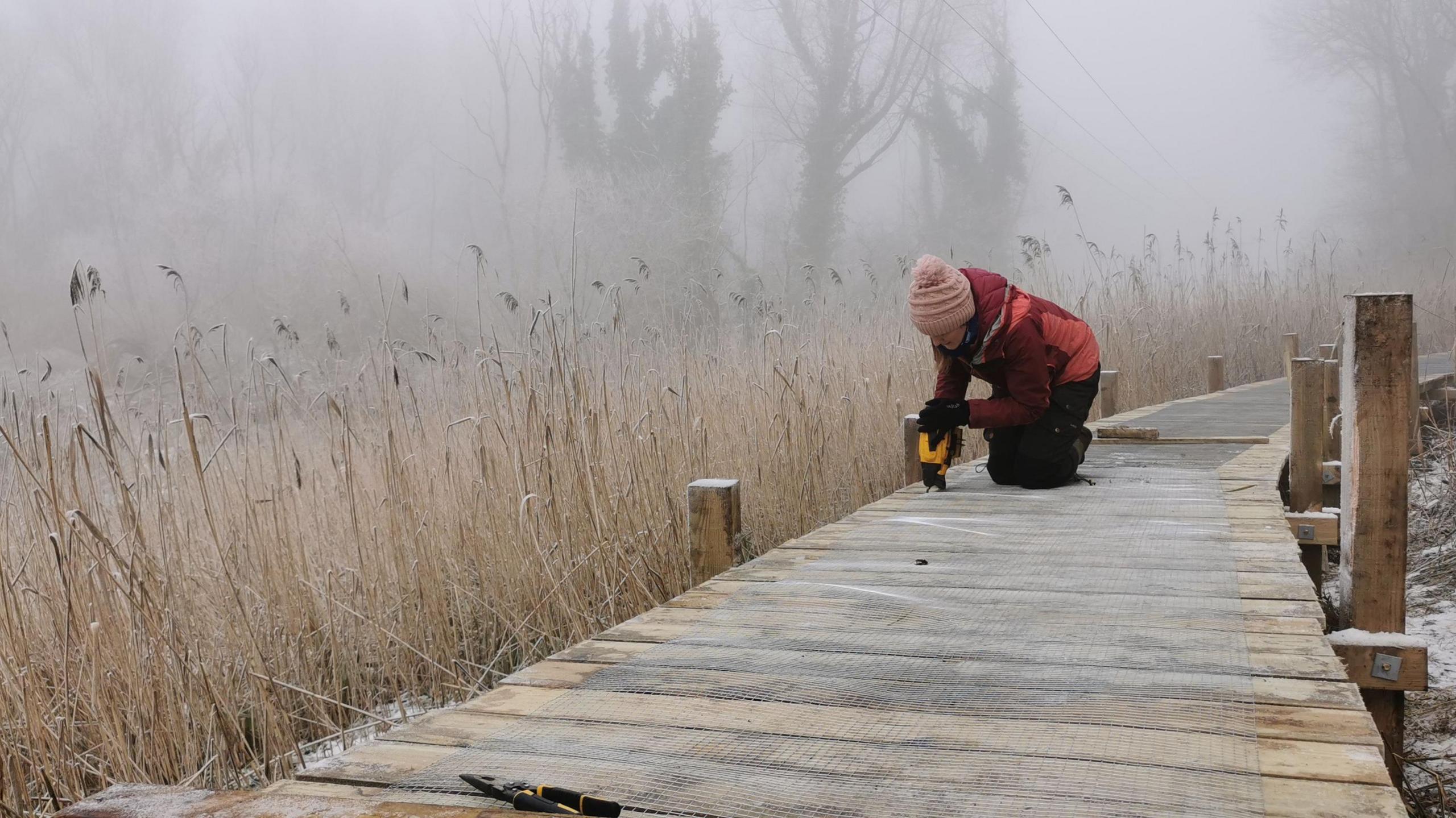 A woman dressed in warm clothes and a woolly hat working on the Hinksey Heights Nature Trail. There is a pair of pliers in sight. It is a foggy day.