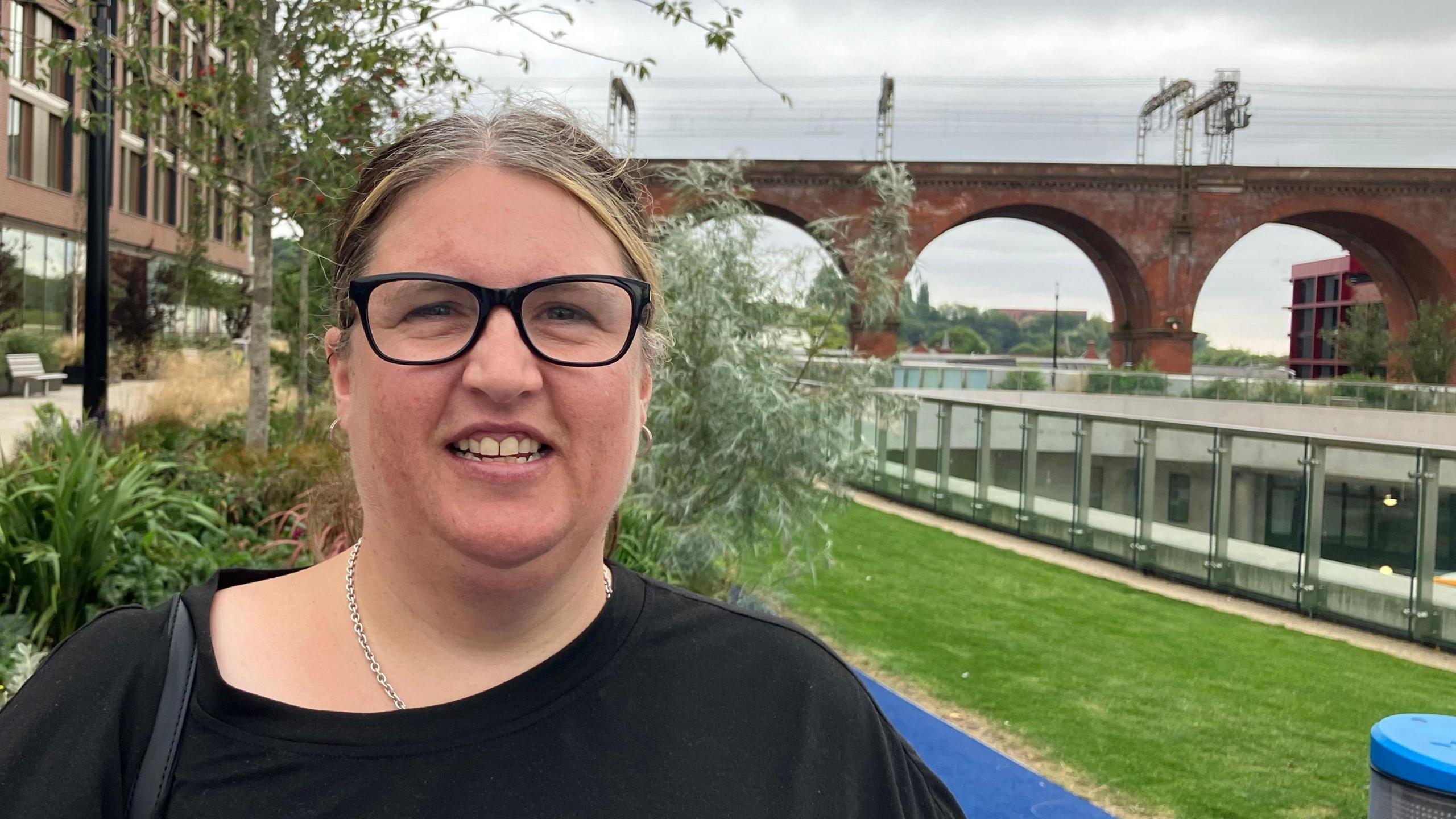 Sarah Williams, who is wearing black glasses and a black t-shirt, smiles while standing close to some young trees and a strip of grass. Behind her, the arches of Stockport's viaduct straddle the view on a grey day.