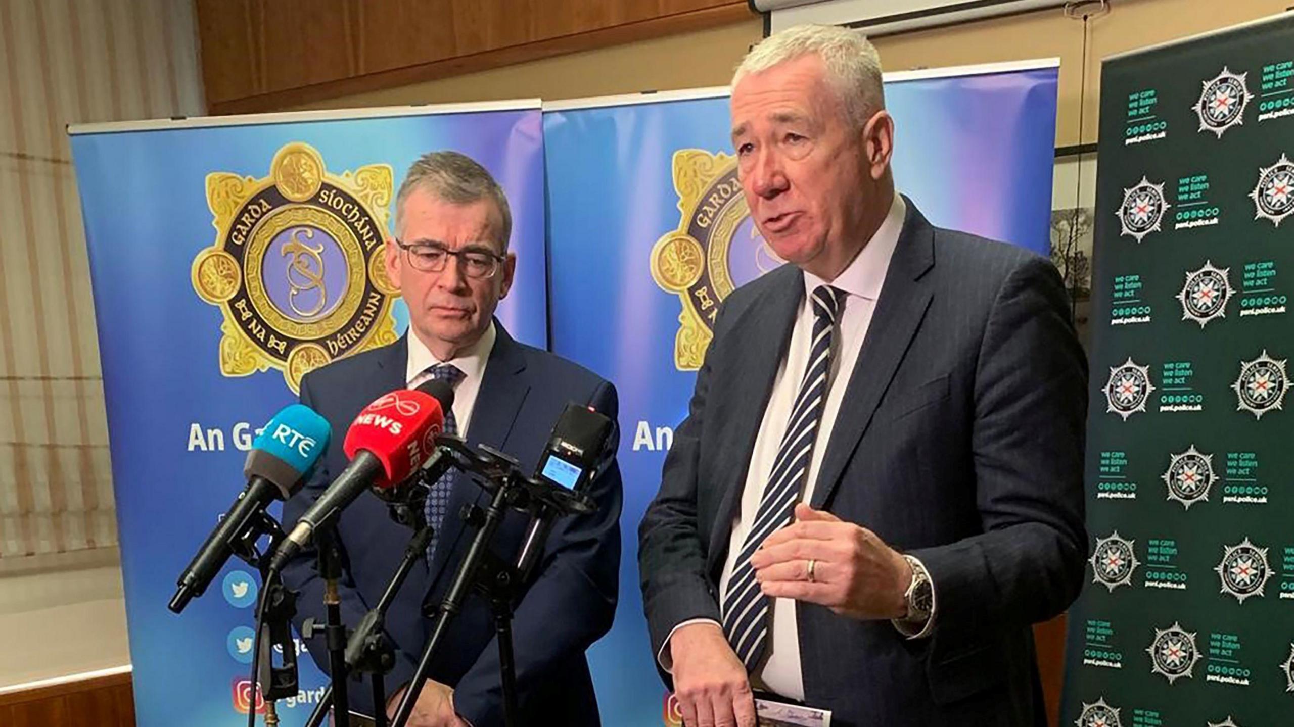 Two men stand in front of several microphones. Behind the man on the left, who is shorter and wearing glasses, there is An Garda Síochána branding. Behind the man on the right, there is PSNI branding. 