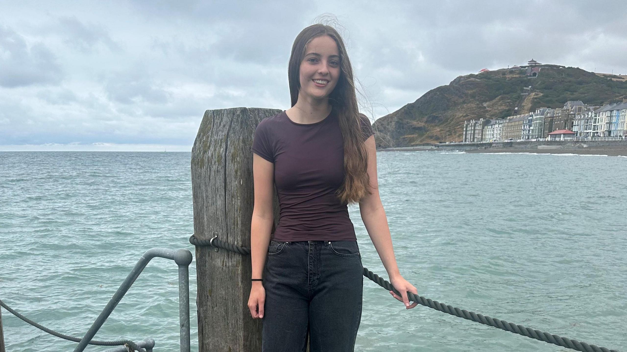 Cadi yn gwisgo crys-t brown a jeans du yn sefyll ar lanfa yn pwyso ar raff gyda'r mor a ffrynt Aberystwyth yn y cefndir