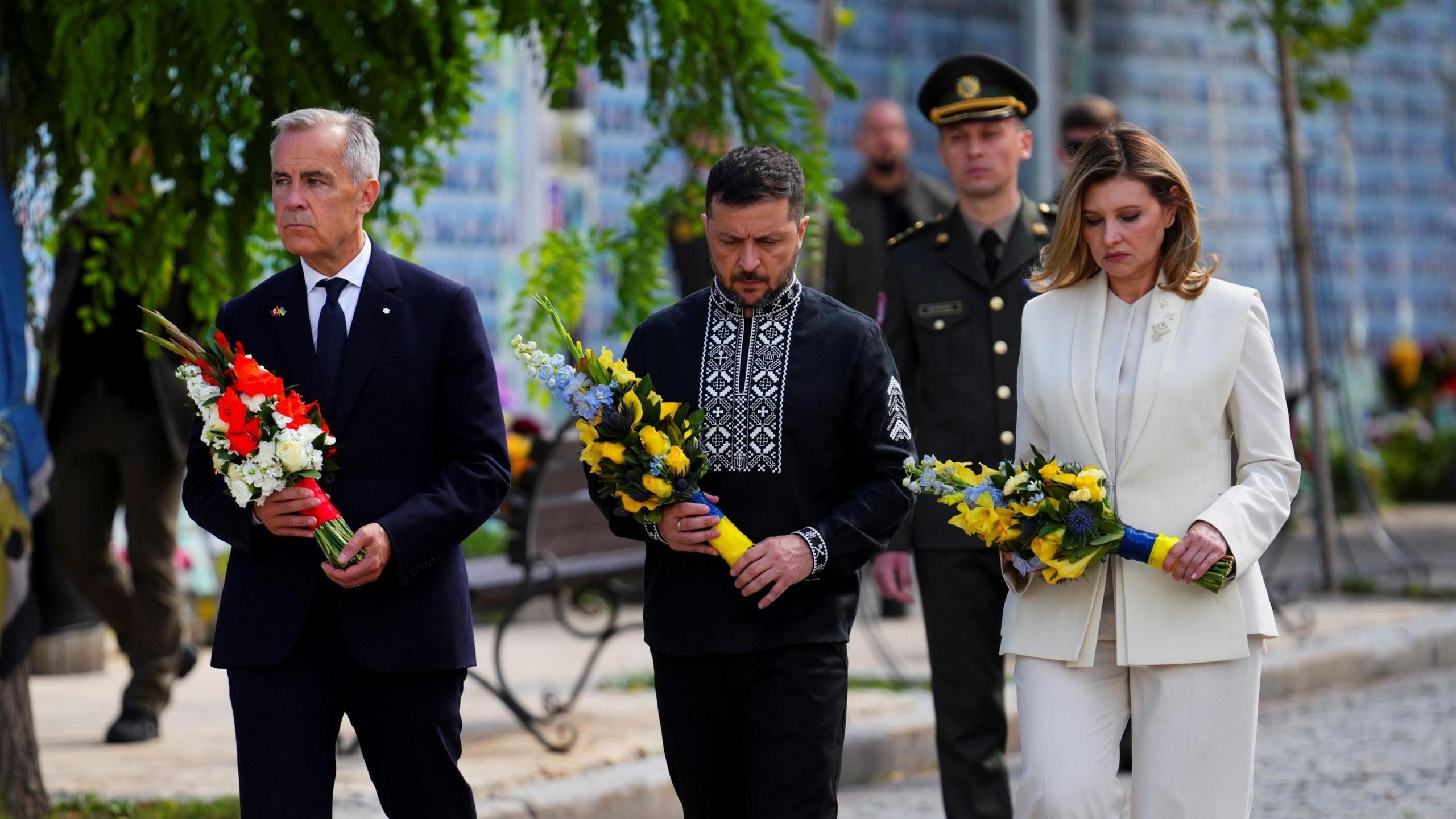 Canada's Prime Minister Mark Carney, his grey hair short and face sombre, holds red and white flowers as he walks in a dark suit on a street in Kyiv, Ukraine. Ukrainian President Volodymyr Zelensky, his hair and beard dark and short, wears a black traditional Ukrainian outfit with white embroidery around the collar and in a line down the front. His wife, Olena Zelenska, wears a white suit. The couple wear sombre expressions and both carry yellow and blue flowers. A soldier walks in the background. 