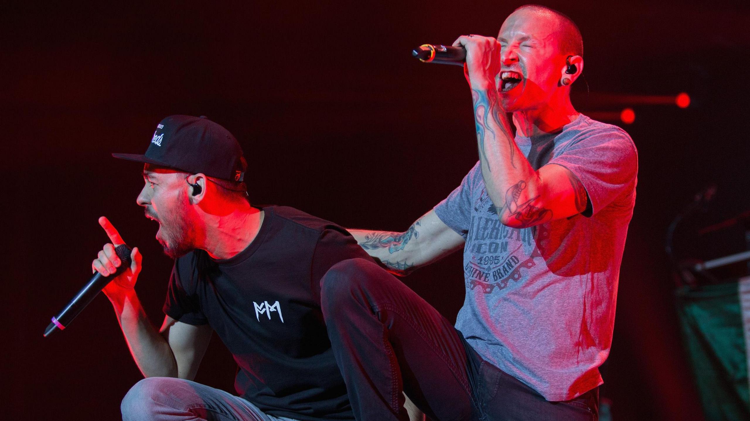 Mike Shinoda and Chester Bennington performing in 2015. They both sing, Chester with his hand on Mike's back as they lean forward off the stage. Mile wears a black cap and T-shirt, Chester wears jeans and a grey T-shirt, revealing heavily tattooed arms. 