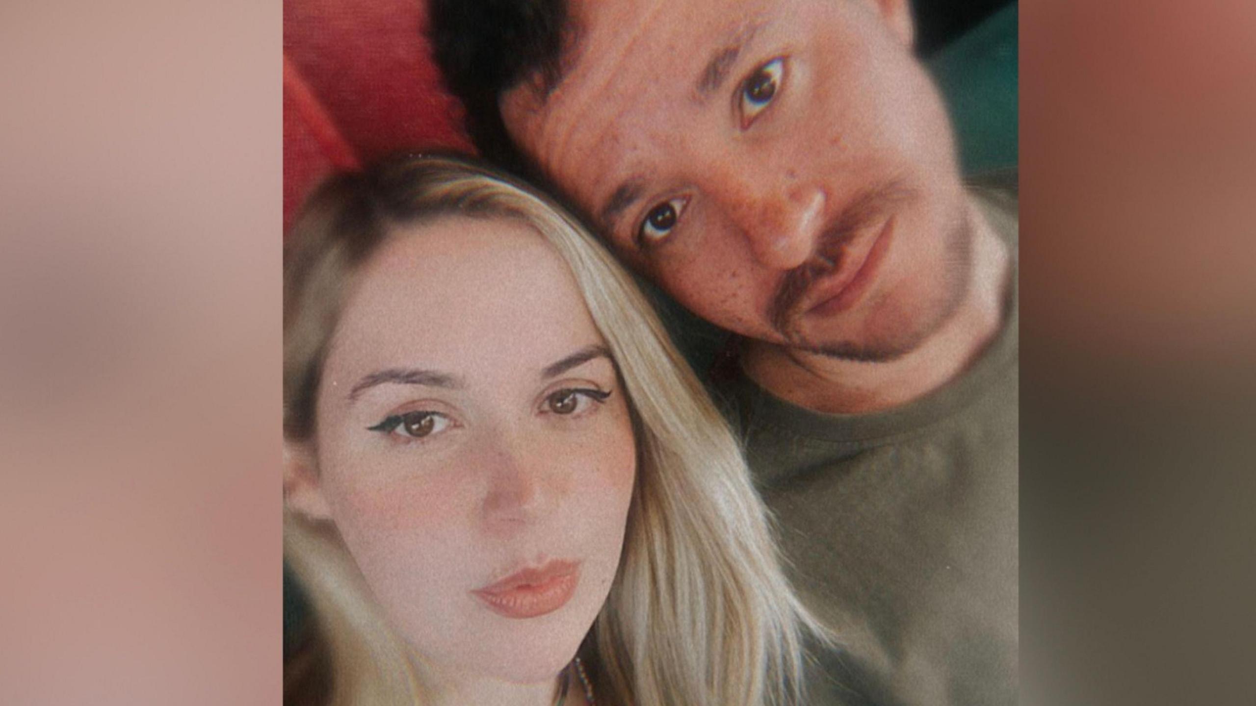 A woman with blonde hair is sat next to a man with moustache and beard and dark hair. They are looking straight at the camera in a selfie pose.