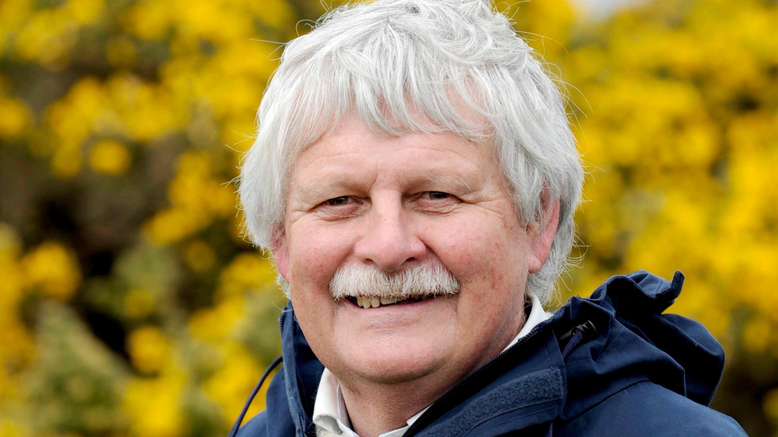 A man with grey hair and a grey moustache in a blue jacket stands in front of a bright yellow gorse bush