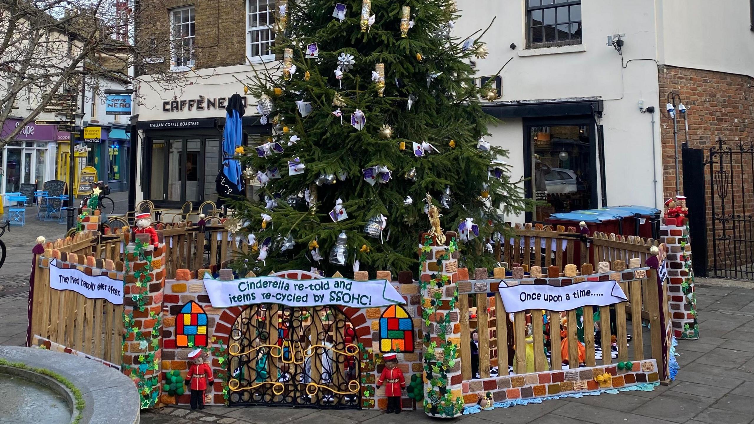 A large Christmas tree in the centre of Hertford, covered in handmade Christmas decorations. A fence surrounds it at the bottom. You can see shops behind the tree and a concrete path. 