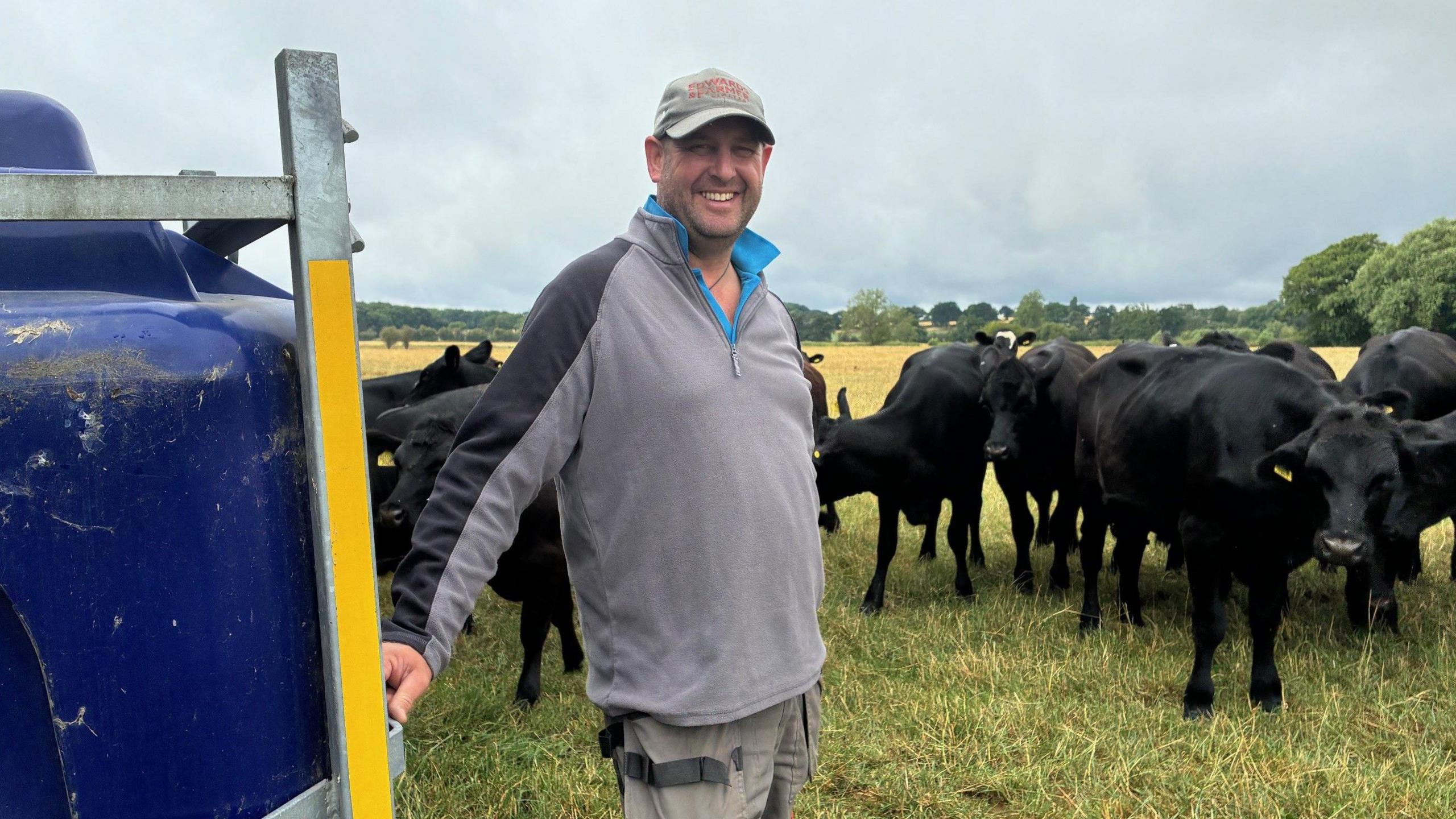 A farmer wearing a grey top, trousers and cap, is standing in a field, next to a blue water tank. Around a dozen black cows are standing in the background.  