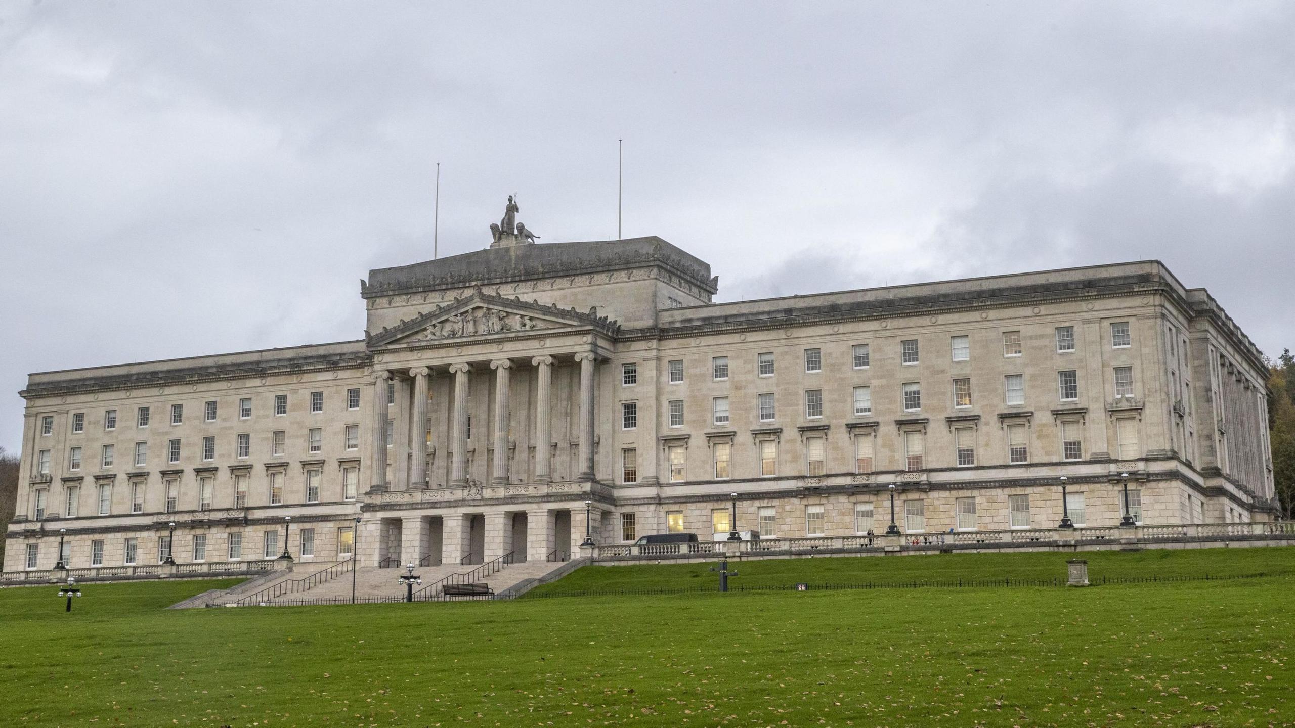 The exterior of Stormont buildings. It shows a large white building sitting on top of a hill. There are six large columns on the outside of the building and a number of windows. The sky is cloudy.