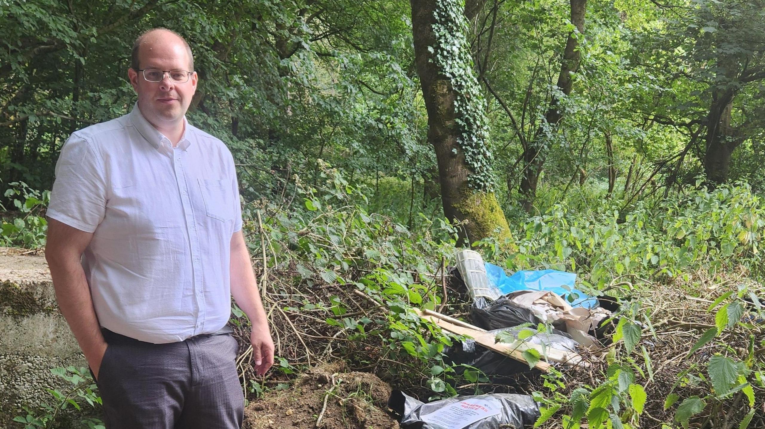 A man wearing grey trousers and a short-sleeve shirt standing next to dumped waste in woodland. He has glasses and balding light brown hair. His right hand is in his right pocket and he is facing the camera.