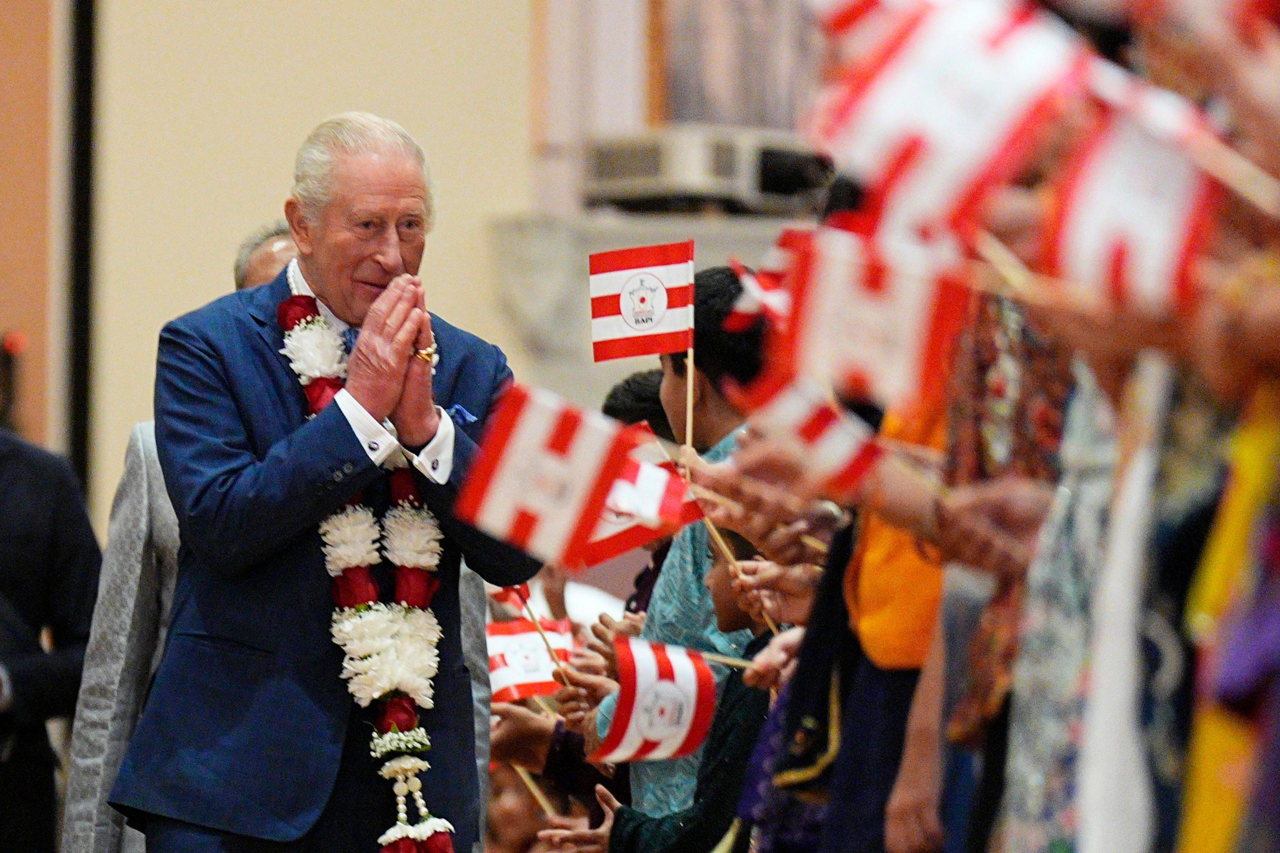 The King clasps his hands together in greeting and smiles as he walks past children holding red and white flags. Children's hands can be seen, but not their faces. He is wearing a dark blue suit and has a red and white garland around his neck.