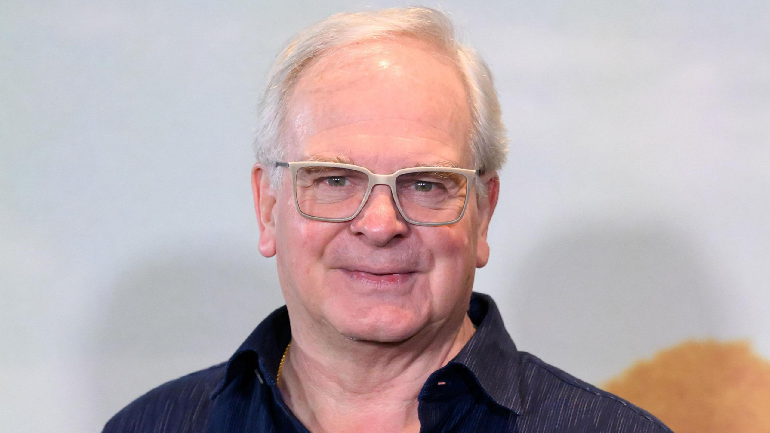 Headshot of Cornwell. He wears a navy shirt, has short white hair, and thick grey framed glasses. 
