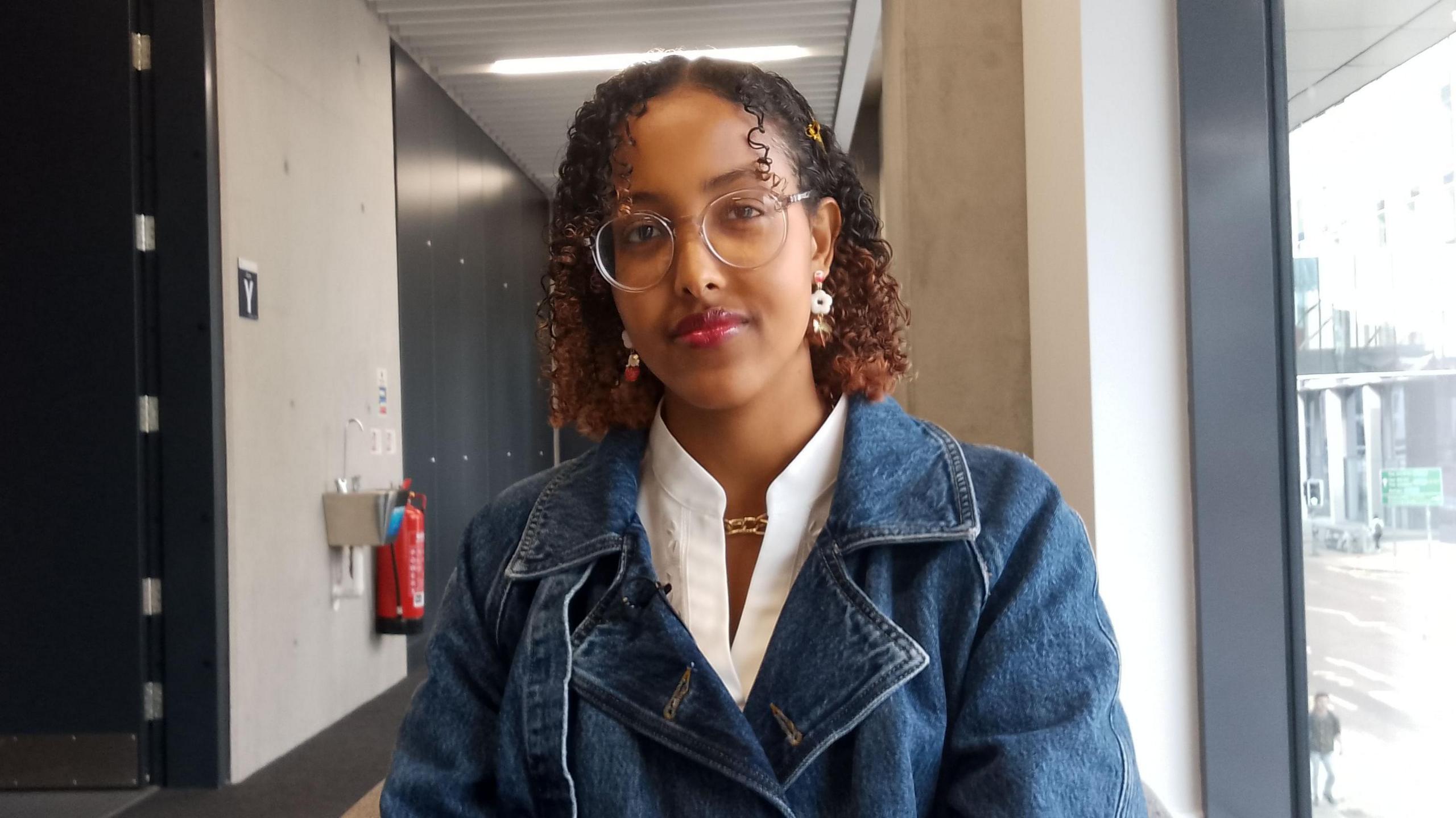 A woman with circular glasses and dark curly hair looks at the camera and smiles